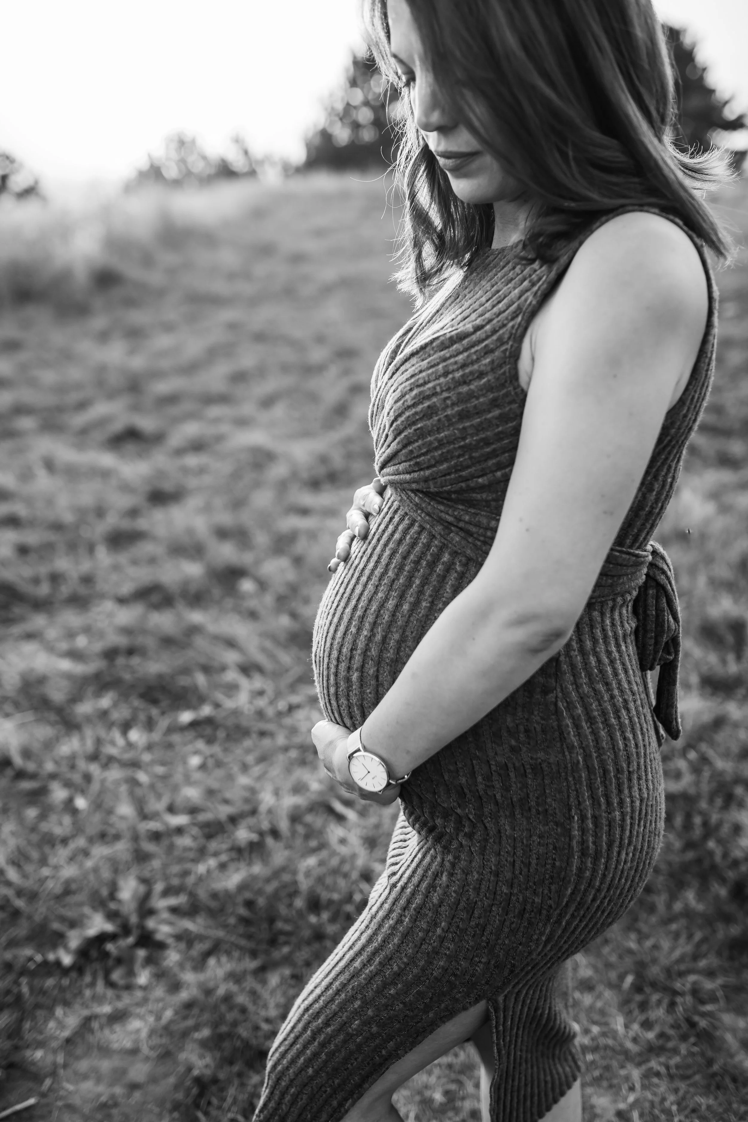 Black and white photo of a pregnant woman standing outdoors, gently holding her belly with both hands, wearing a sleeveless ribbed dress, with her hair falling over her face, in a natural setting with blurred background.