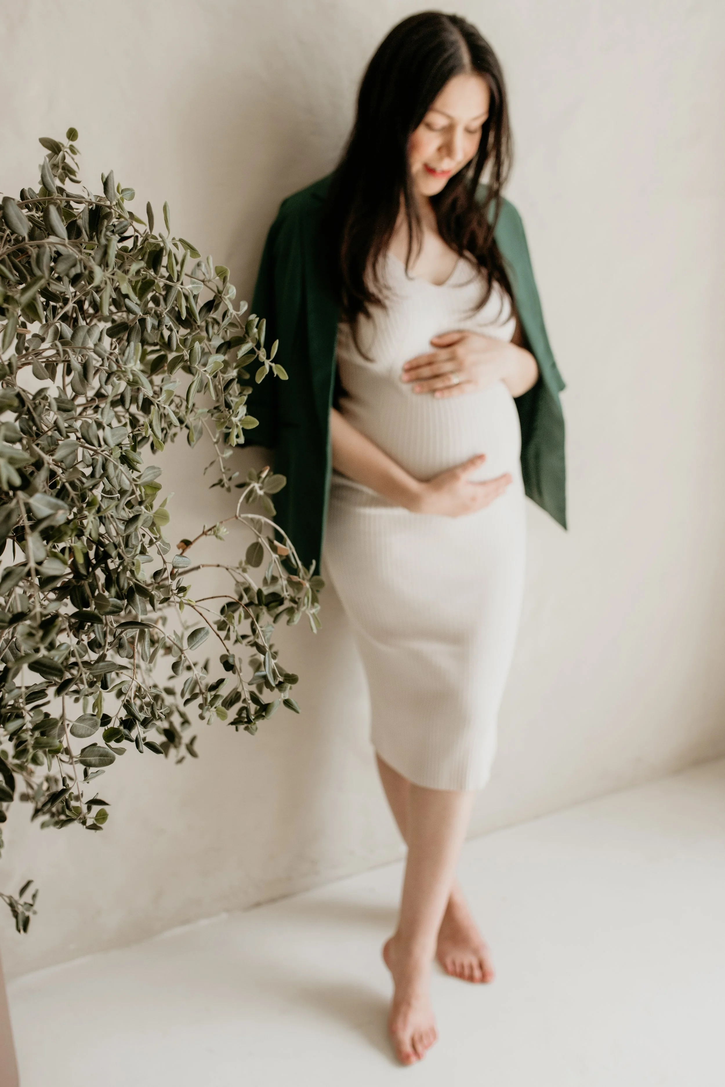 A pregnant woman with long dark hair, wearing a form-fitting white dress and a green jacket, stands barefoot against a beige wall. She is looking down with a gentle smile and resting her hands on her belly.