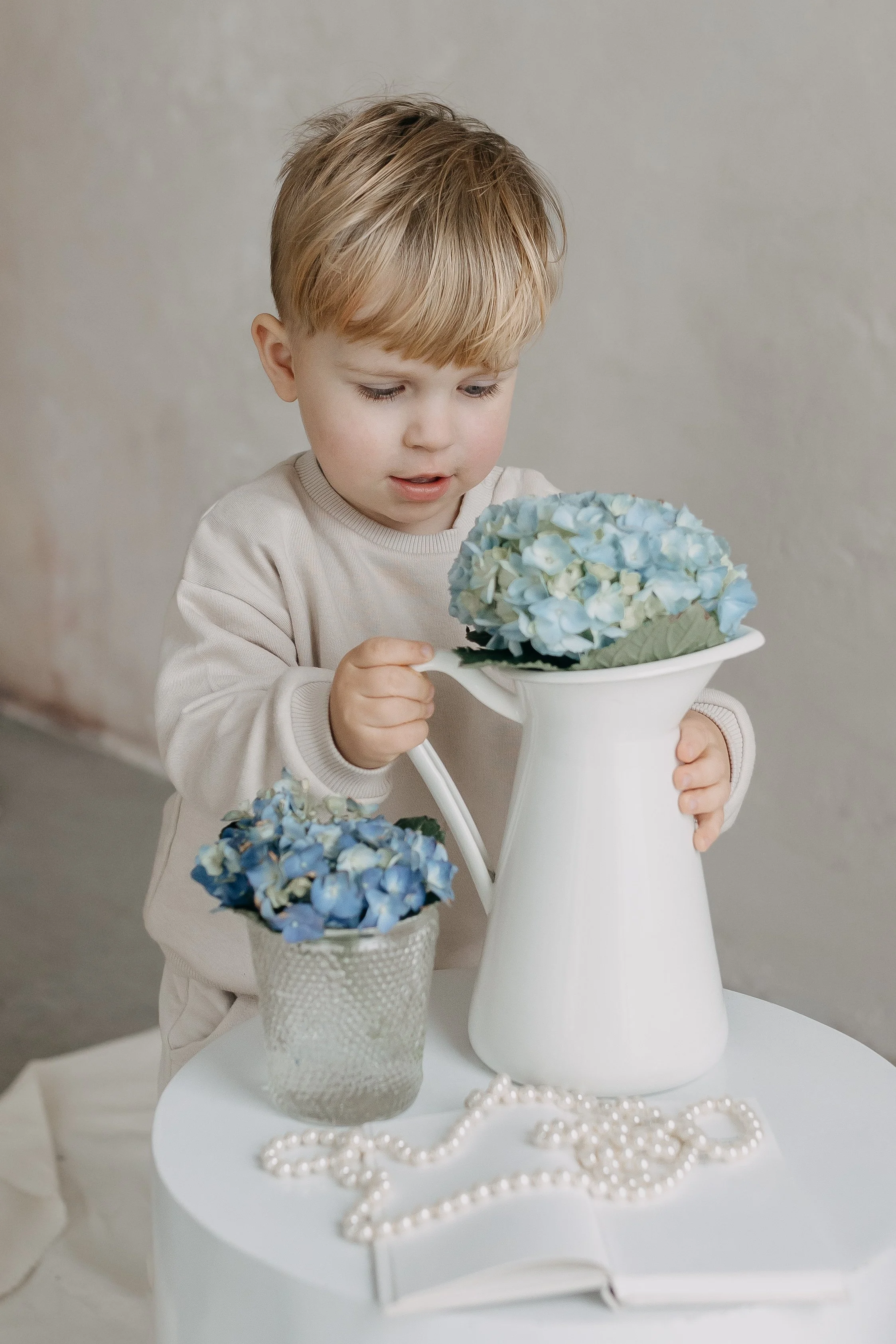 A young boy with blond hair looking at a bouquet of blue hydrangeas in a white vase, with pearls on a white table in front of him.