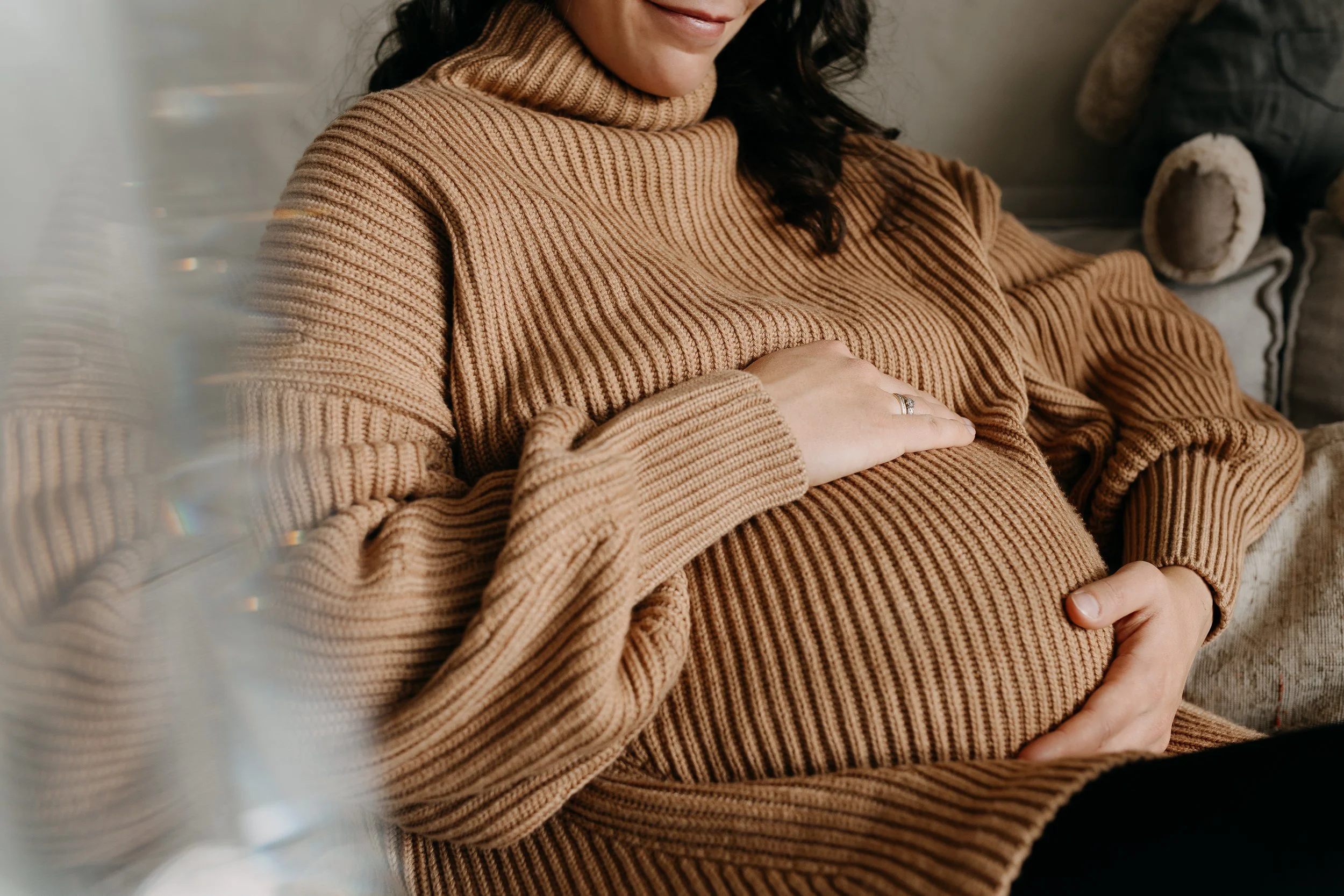 A woman with dark, curly hair wearing a tan, ribbed turtleneck sweater, smiling gently and resting her hand on her belly, which is covered by the same sweater. She is sitting on a soft surface, and there are cozy blankets nearby.