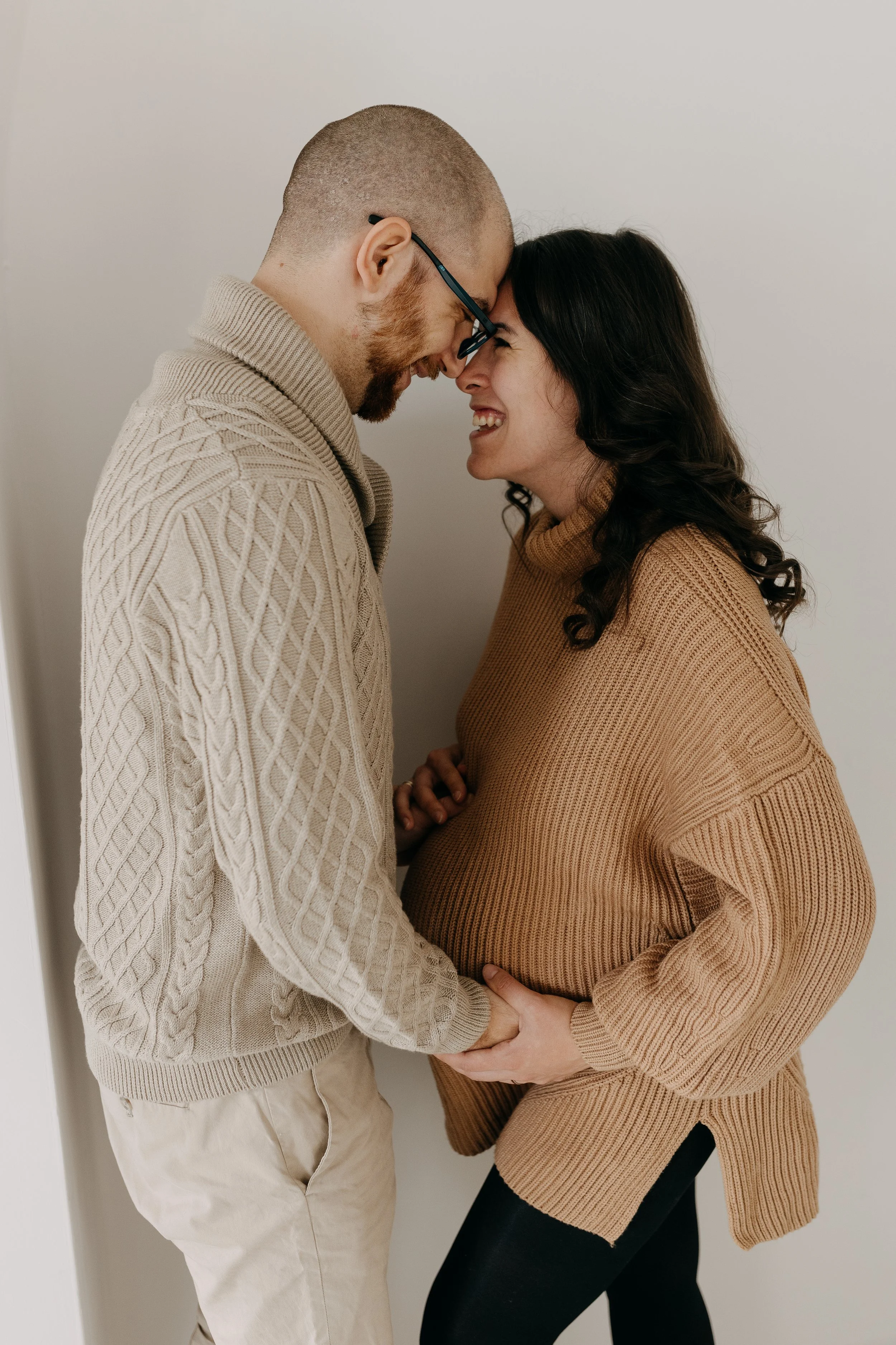 A couple is standing close with their foreheads touching, smiling, and holding hands. The woman appears pregnant.