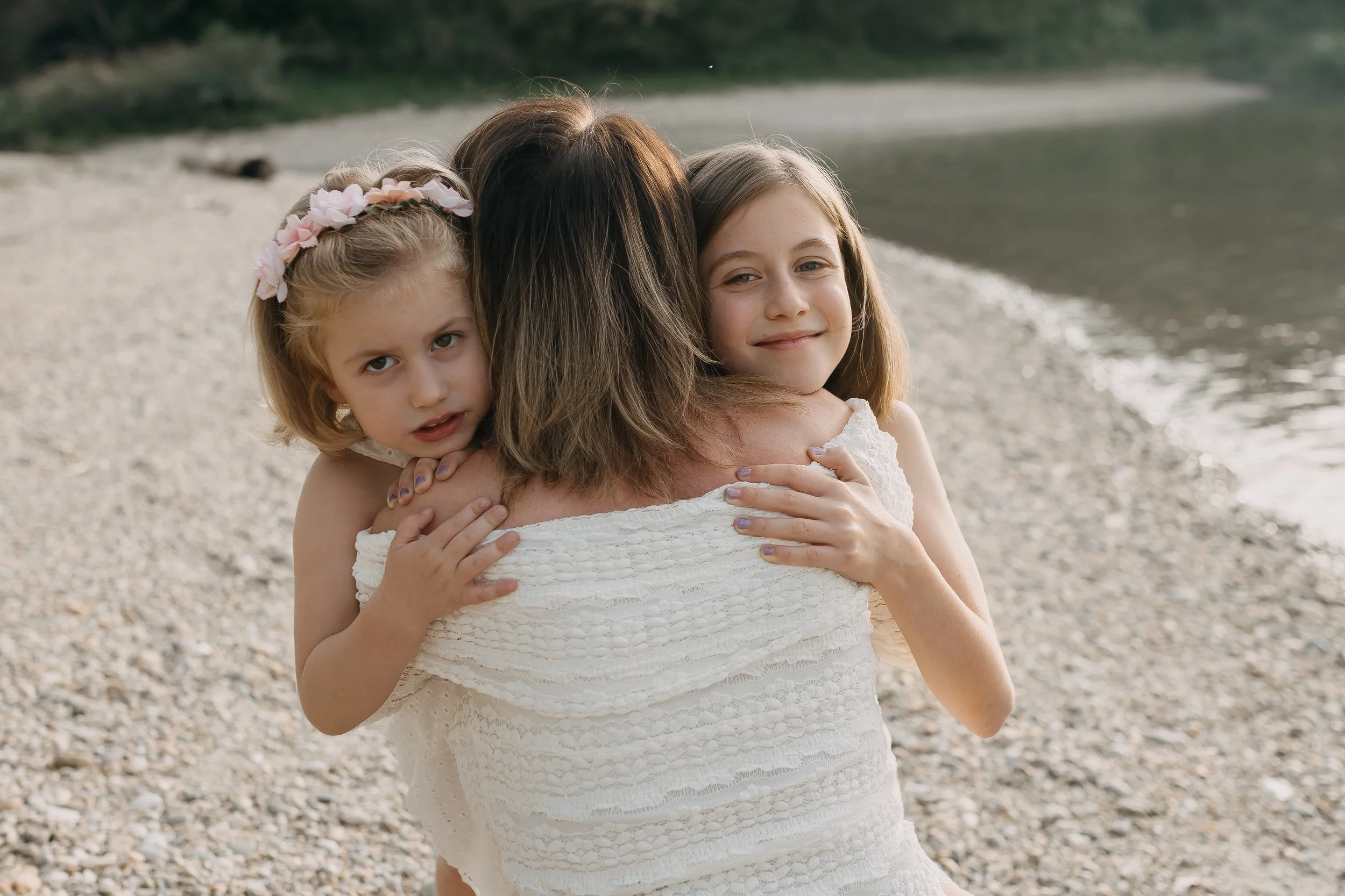 A woman hugging two young girls on a rocky beach with water in the background.
