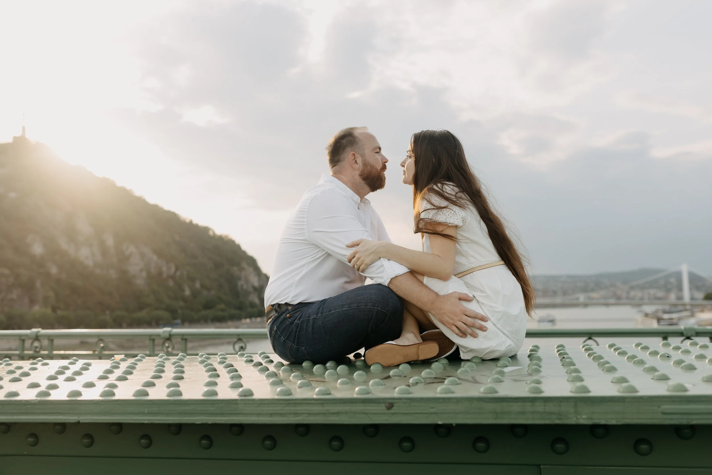 A man and woman are sitting on the ground, facing each other, holding hands and leaning in for a kiss. They are outdoors on a rooftop with small round objects scattered on the surface around them. The background shows a cloudy sky, a hill or mountain