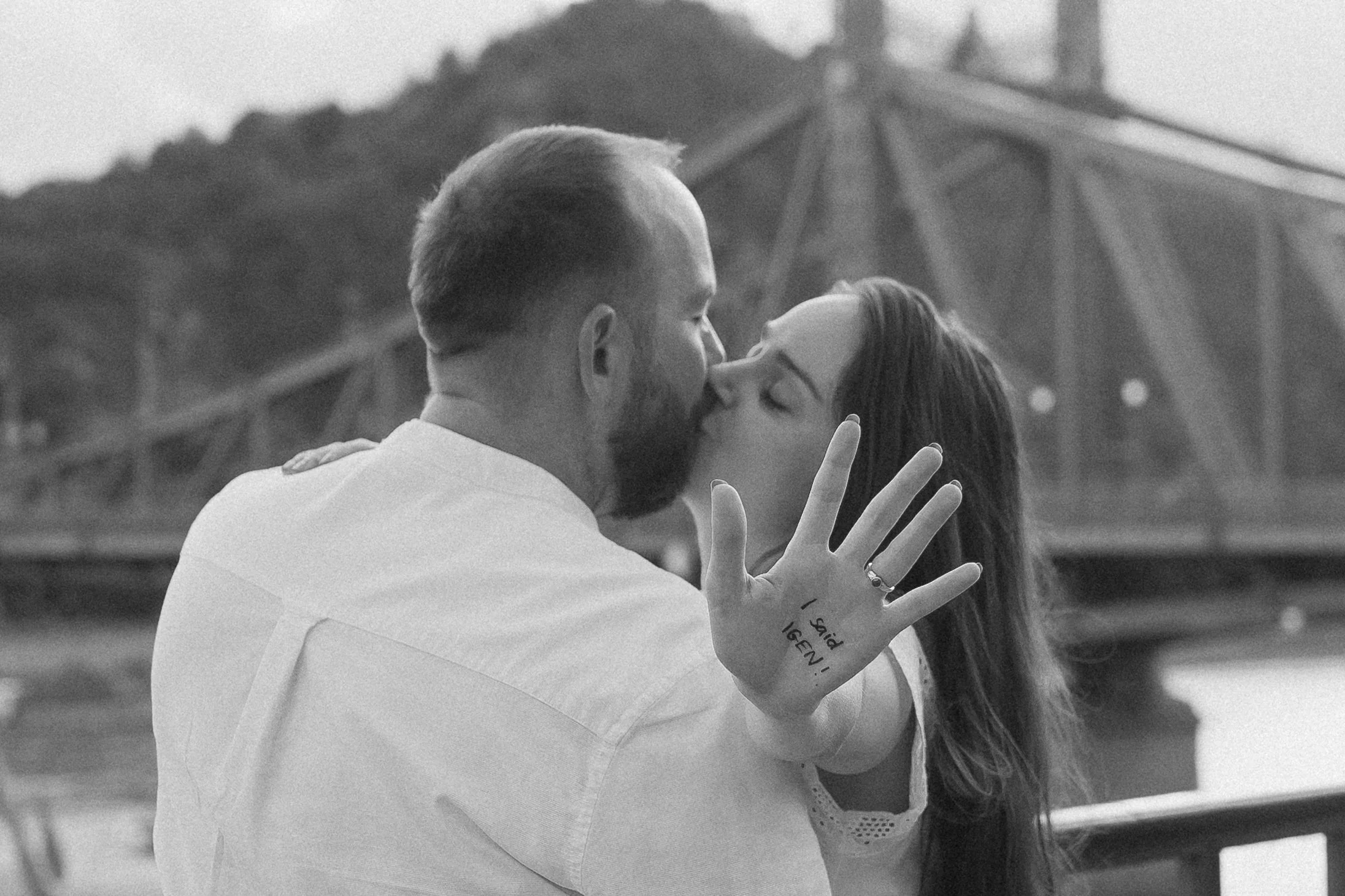 A black-and-white photo of a couple sharing a kiss outdoors, with a bridge in the background, one person's hand visible with words written on it.
