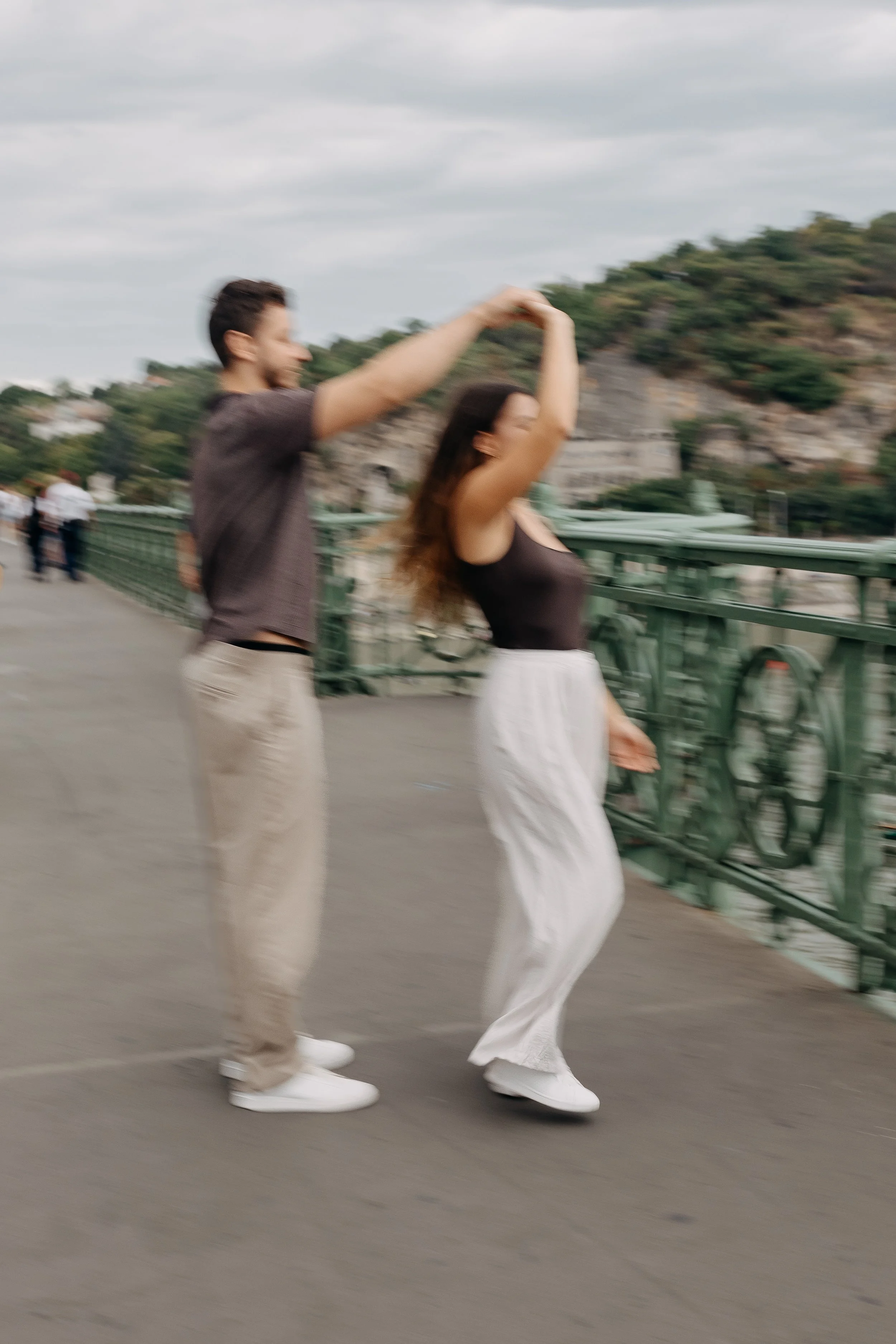 A couple dancing on a sidewalk on a bridge, with a hill and trees in the background and cloudy sky above.