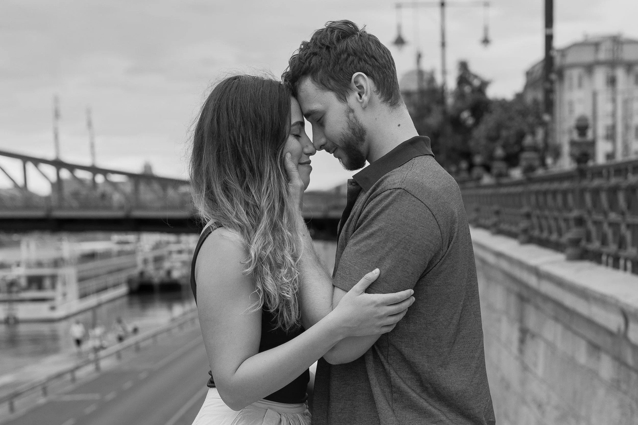 A black and white photo of a man and woman embracing closely with their foreheads touching, outdoors near a bridge and city buildings.