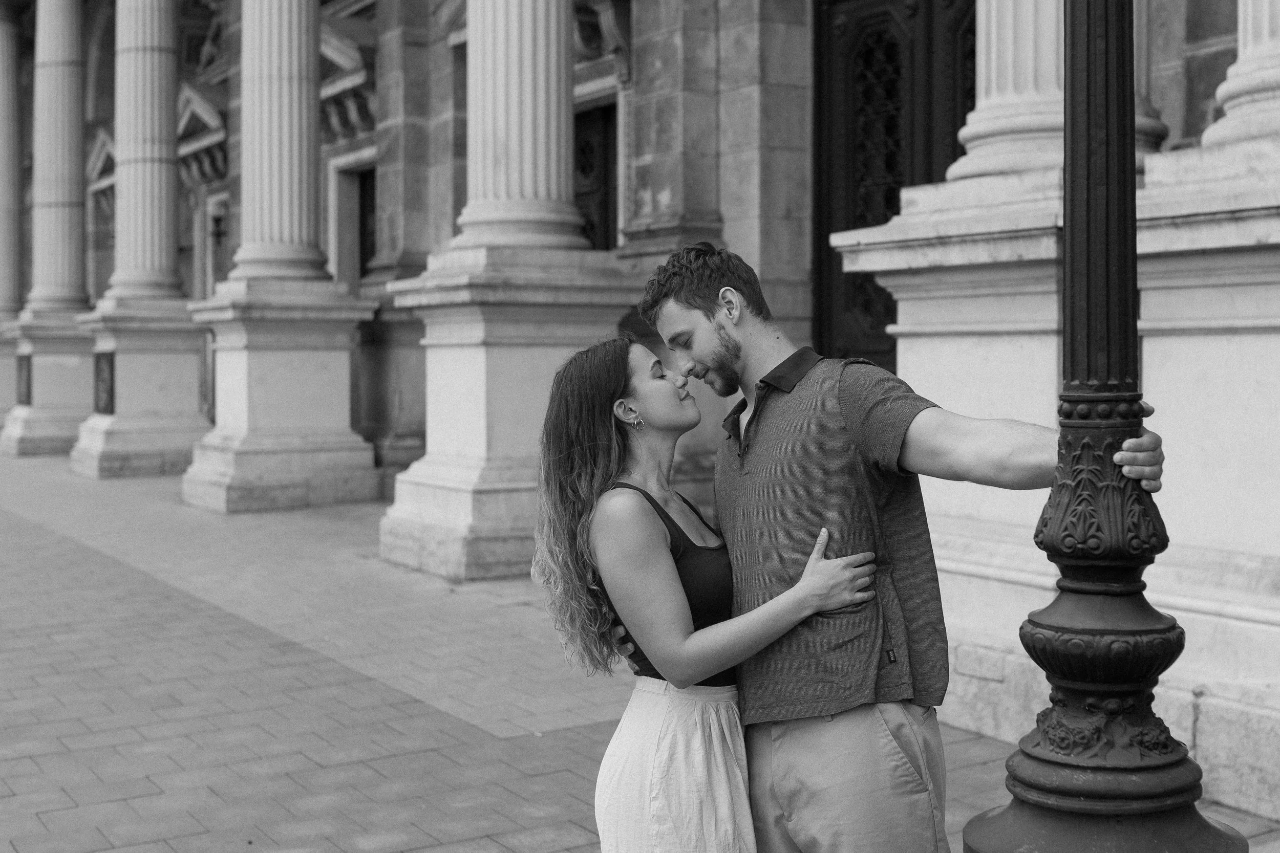 A couple is sharing a tender moment, standing close with their foreheads touching. They are outdoors in front of classical columns and architecture, with the woman holding the man around the waist and the man holding her by the back. The scene is in 
