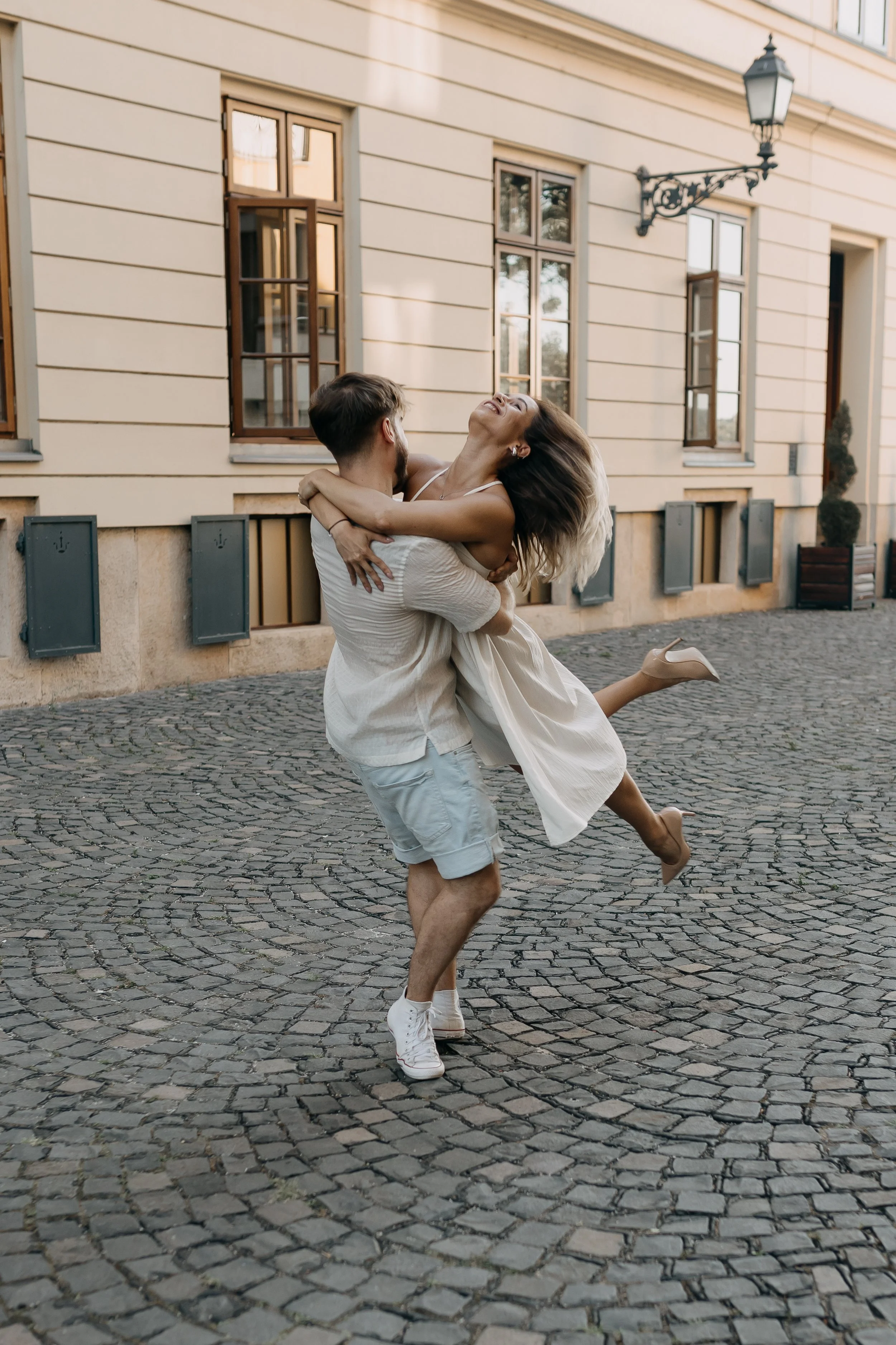 A man is lifting a woman in his arms on a cobblestone street in front of a beige building with tall windows and outdoor wall lanterns. The woman is smiling with her head tilted back and her hair flowing, while the man has his back to the camera. They