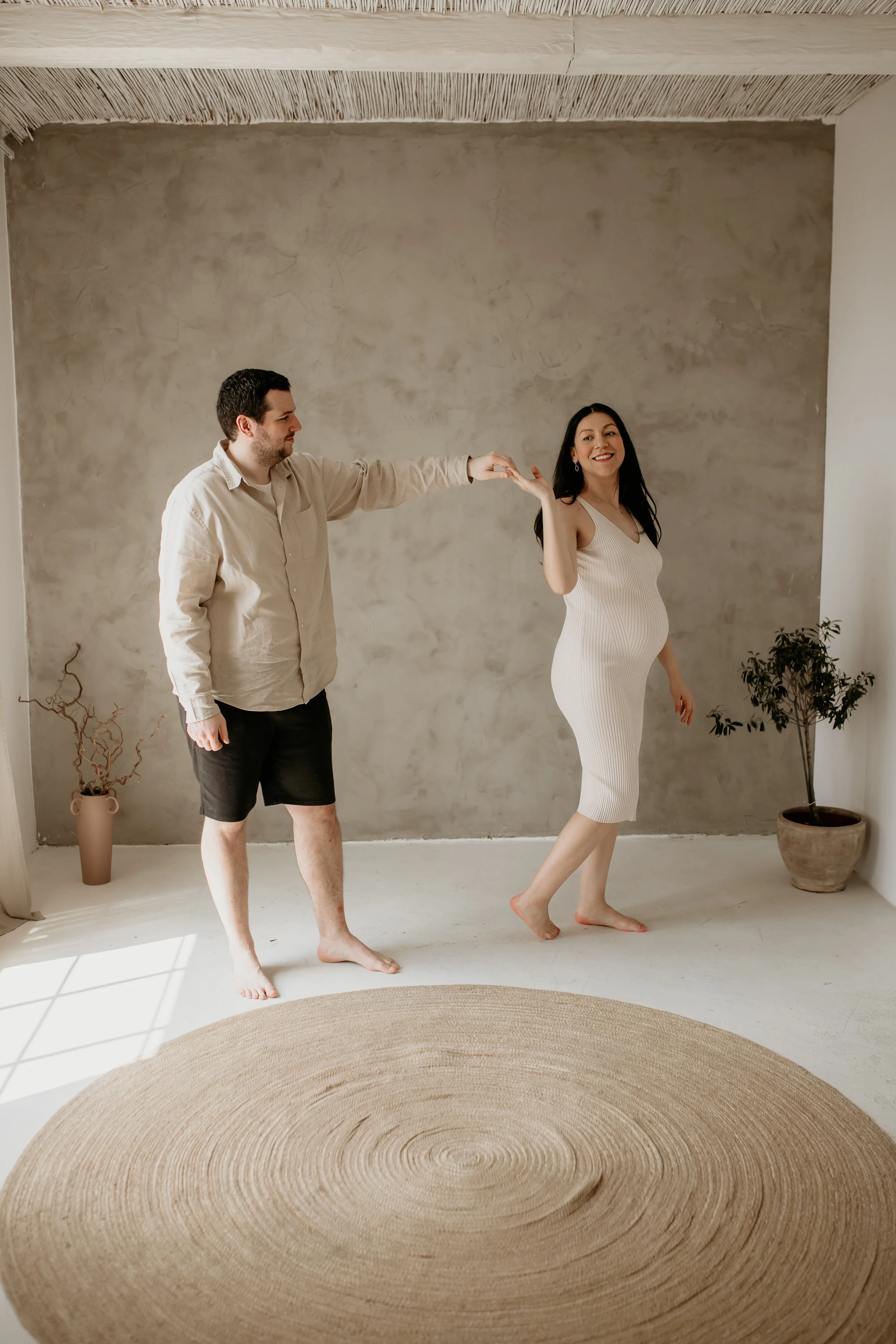 A man and pregnant woman standing barefoot on a plain floor in a room with minimalist decor. The man is reaching out to touch the woman's hand, and she is smiling and looking happy.