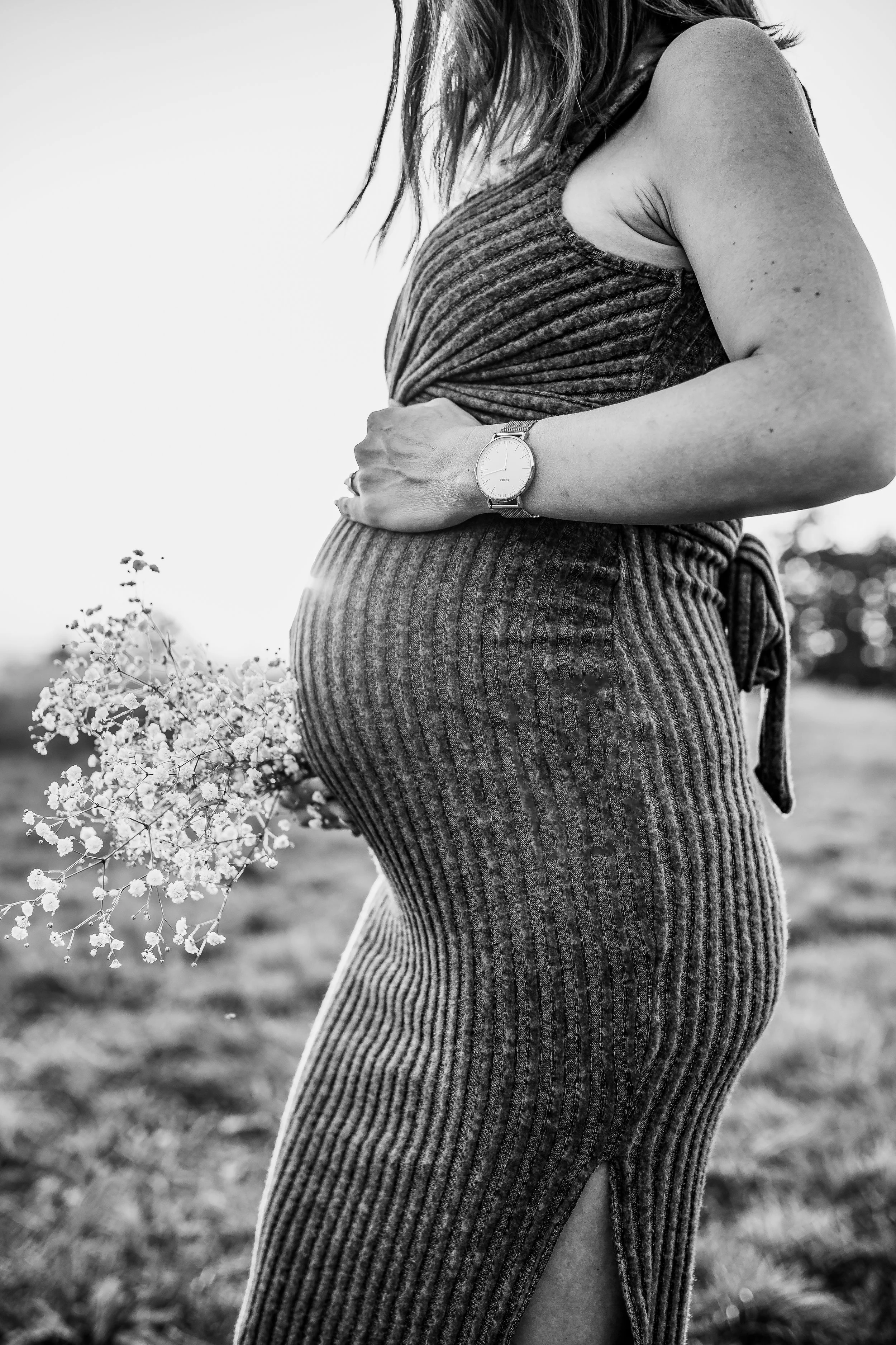 A pregnant woman in a striped dress with a watch on her wrist, standing outdoors near flowers, captured in black and white.