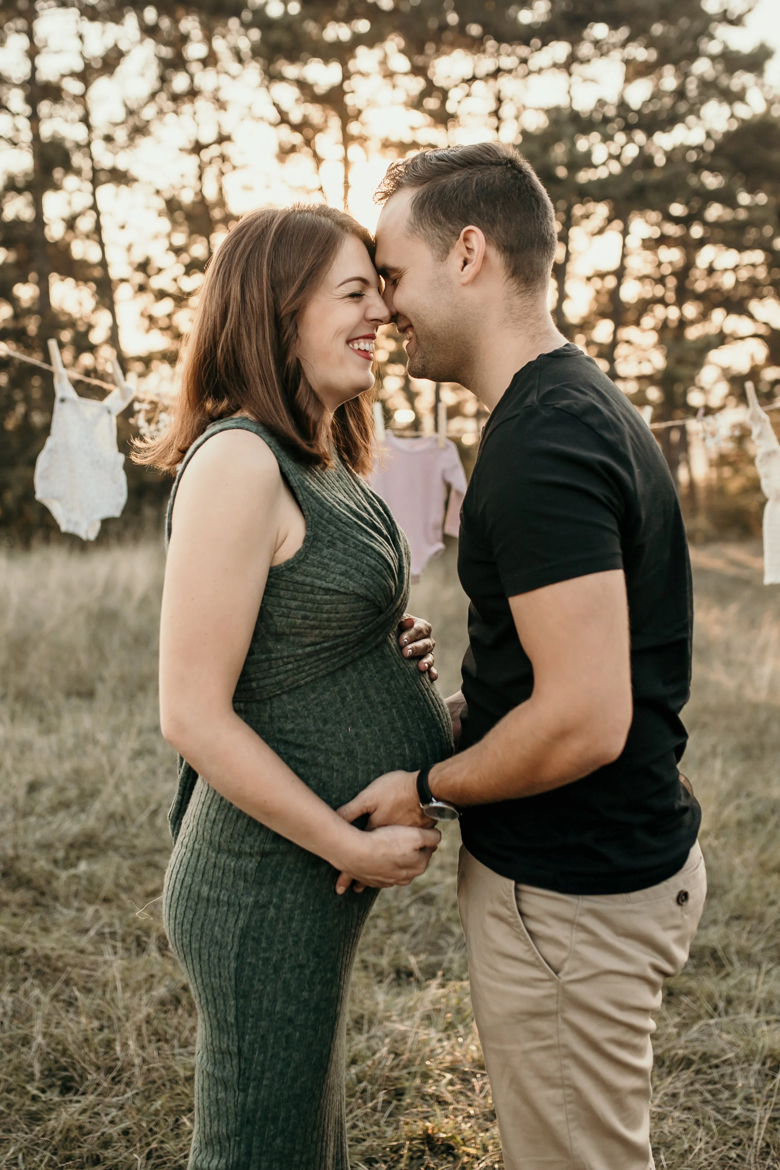 A pregnant woman and a man stand outdoors, touching foreheads and smiling, with trees and hanging laundry in the background, during sunset.