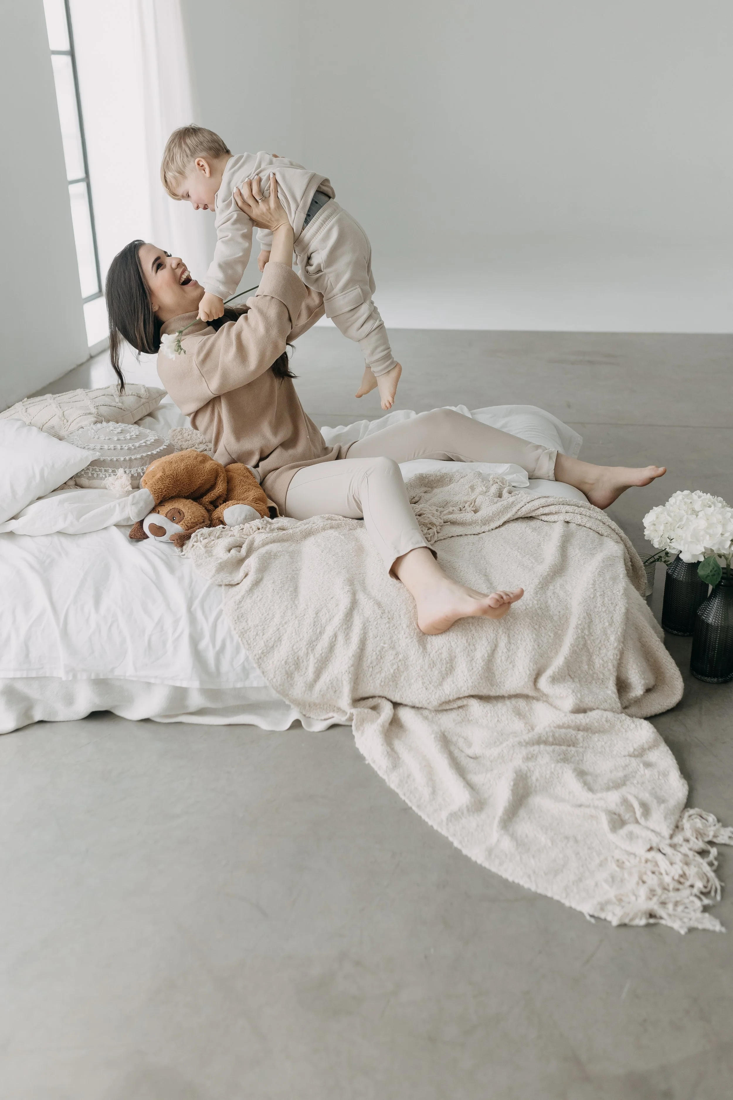 A woman and a young boy playing on a bed in a bright room with large windows, surrounded by soft blankets and plush toys.