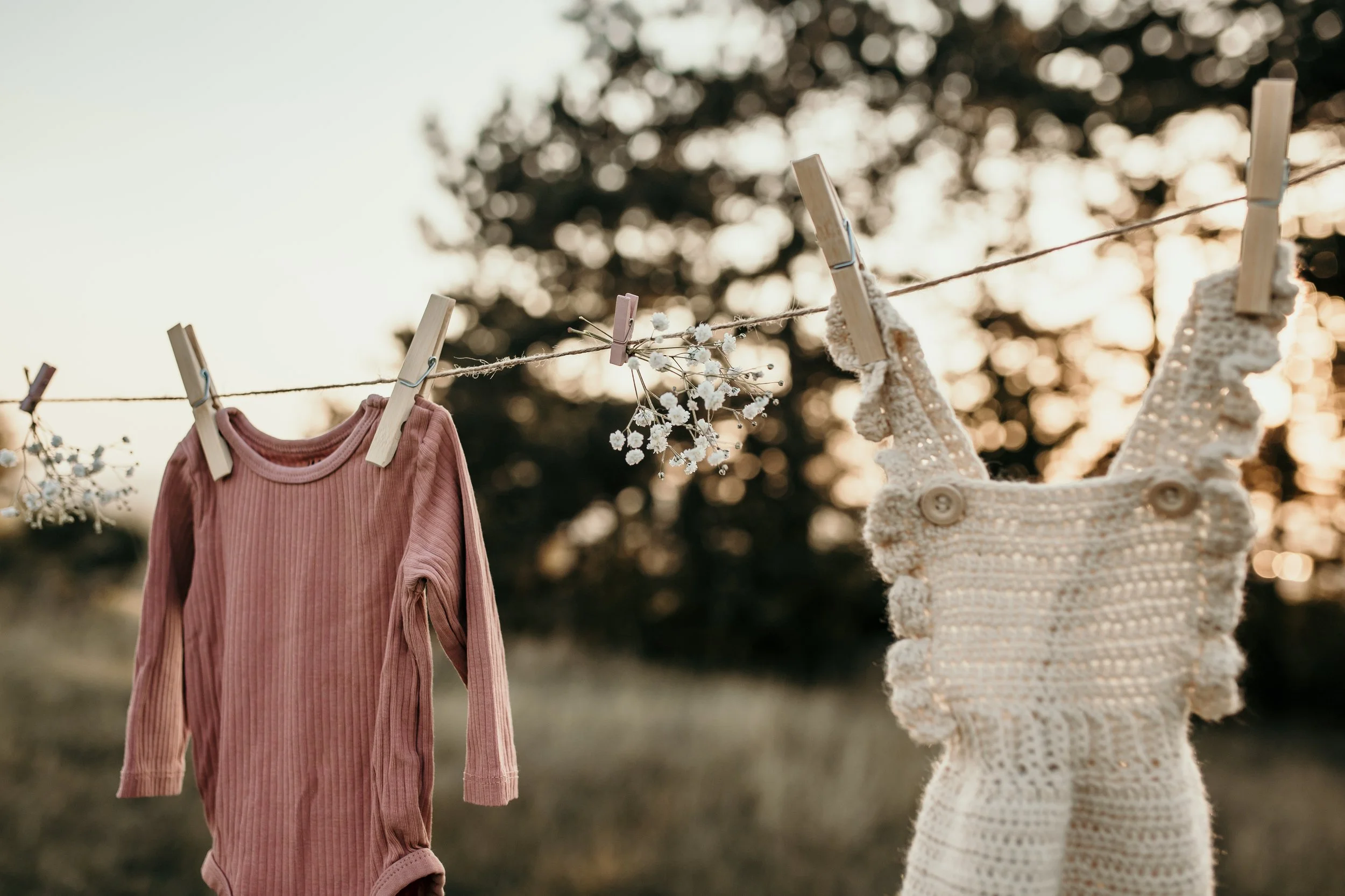 Clothespins hanging on a clothesline with a pink long-sleeve shirt, a bunch of small white flowers, and a knitted baby sweater hanging on a sunny day outdoors.