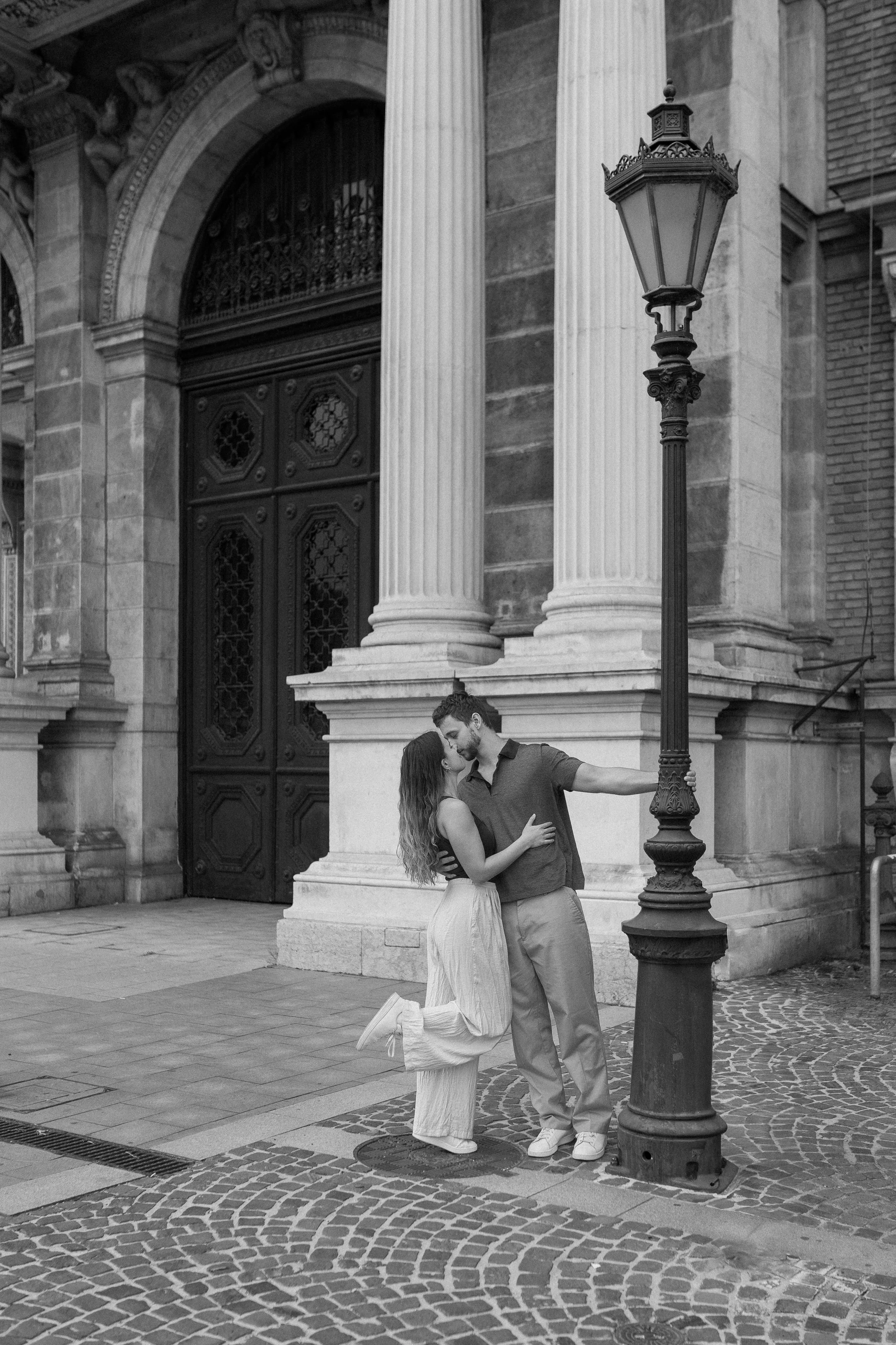 A couple stands close together on a city street at night, posing near a streetlamp, in front of large columns and ornate wooden doors of a historic building.