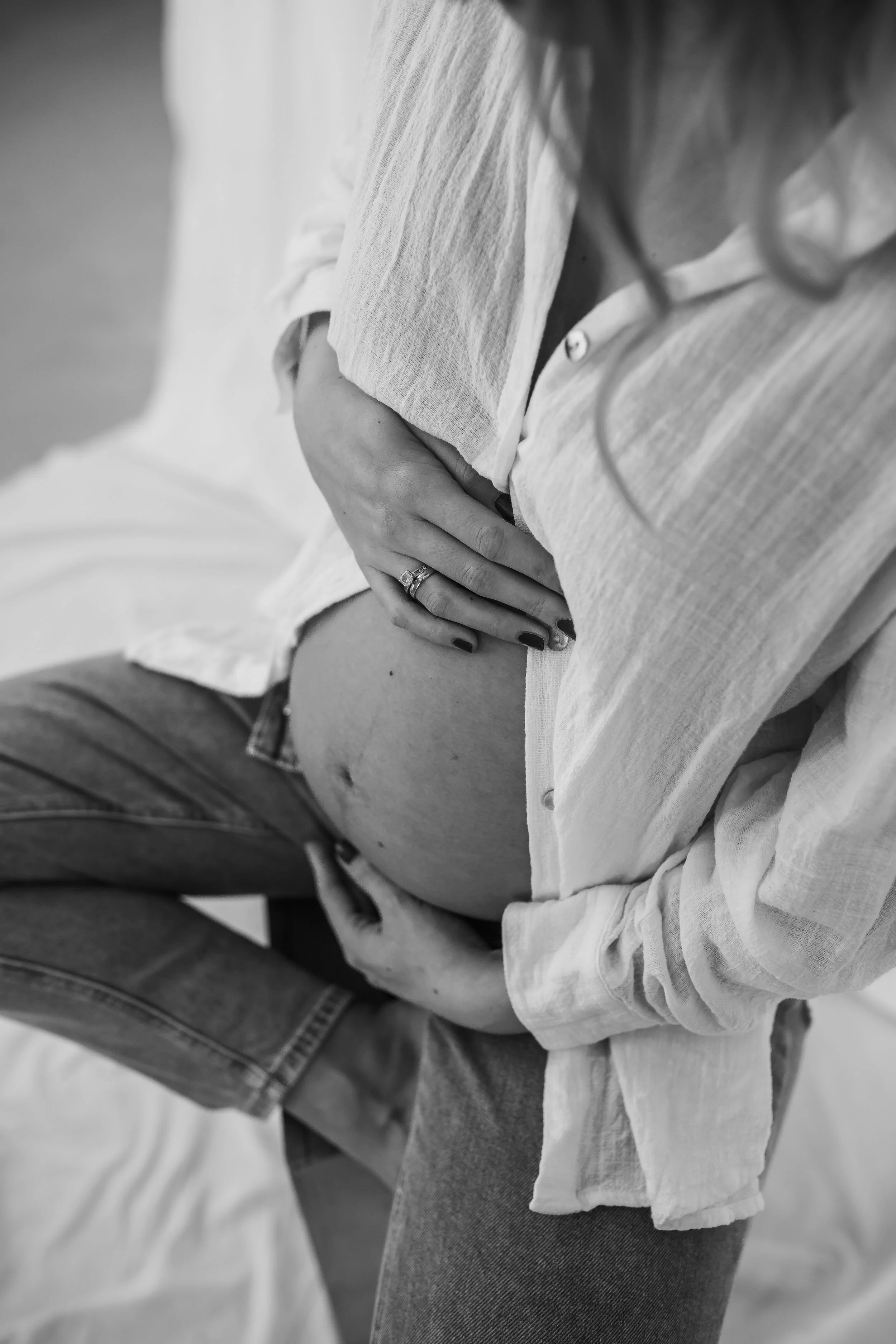 A pregnant woman wearing a white blouse and jeans, with her hand resting on her baby bump. The image is in black and white.