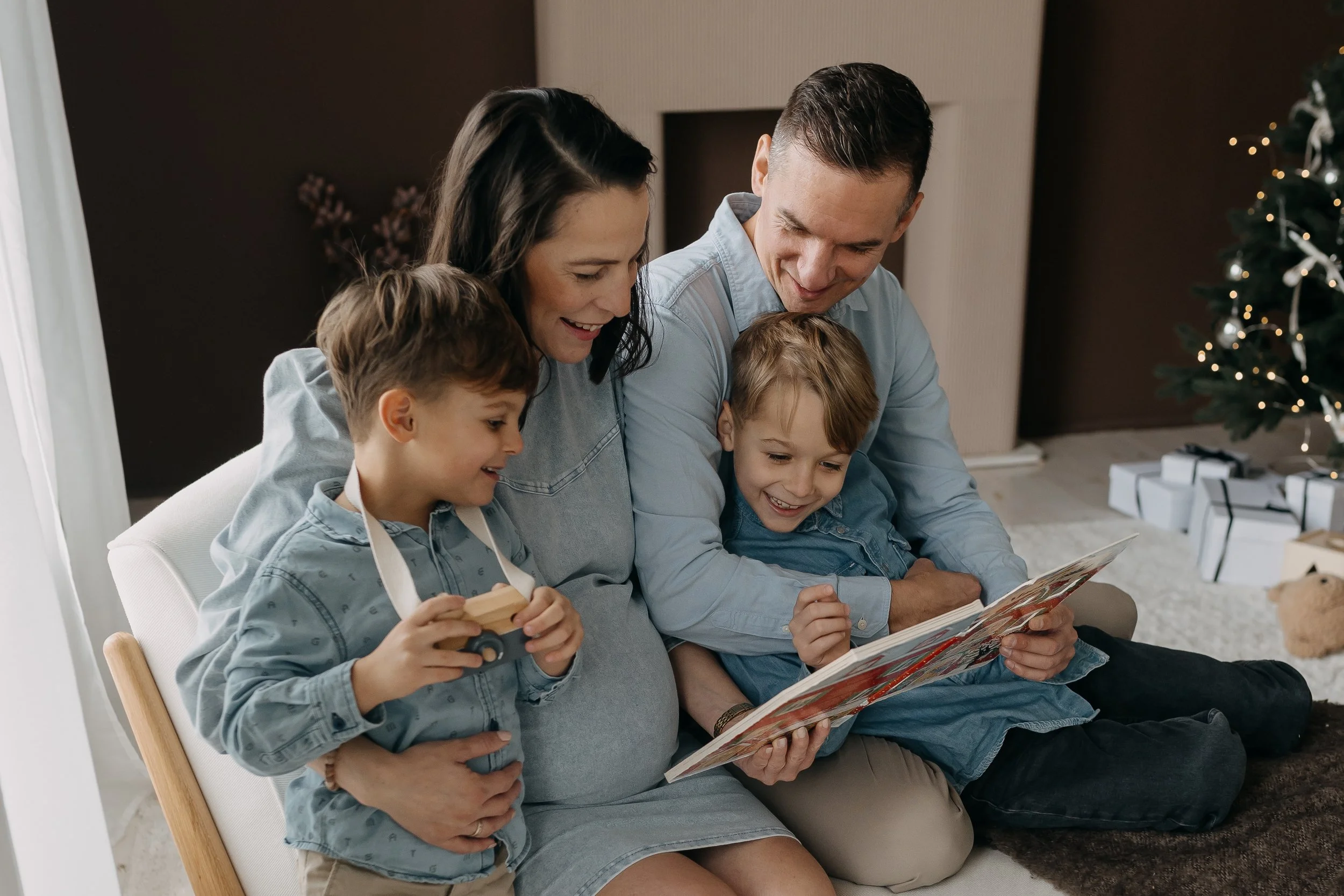 A family of four sitting on a couch in a living room decorated for Christmas, looking at a photograph, with a Christmas tree in the background.