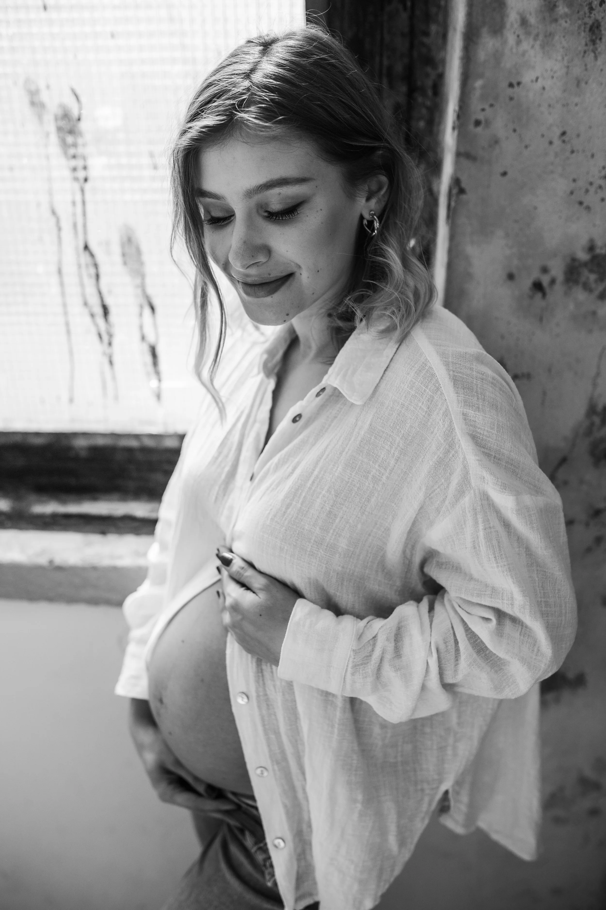 Black and white photo of a pregnant woman with curly hair, smiling softly with her eyes closed. She is wearing a loose, button-up shirt and hoops earrings, standing next to a window with a curtain, against a textured wall. She is cradling her belly.