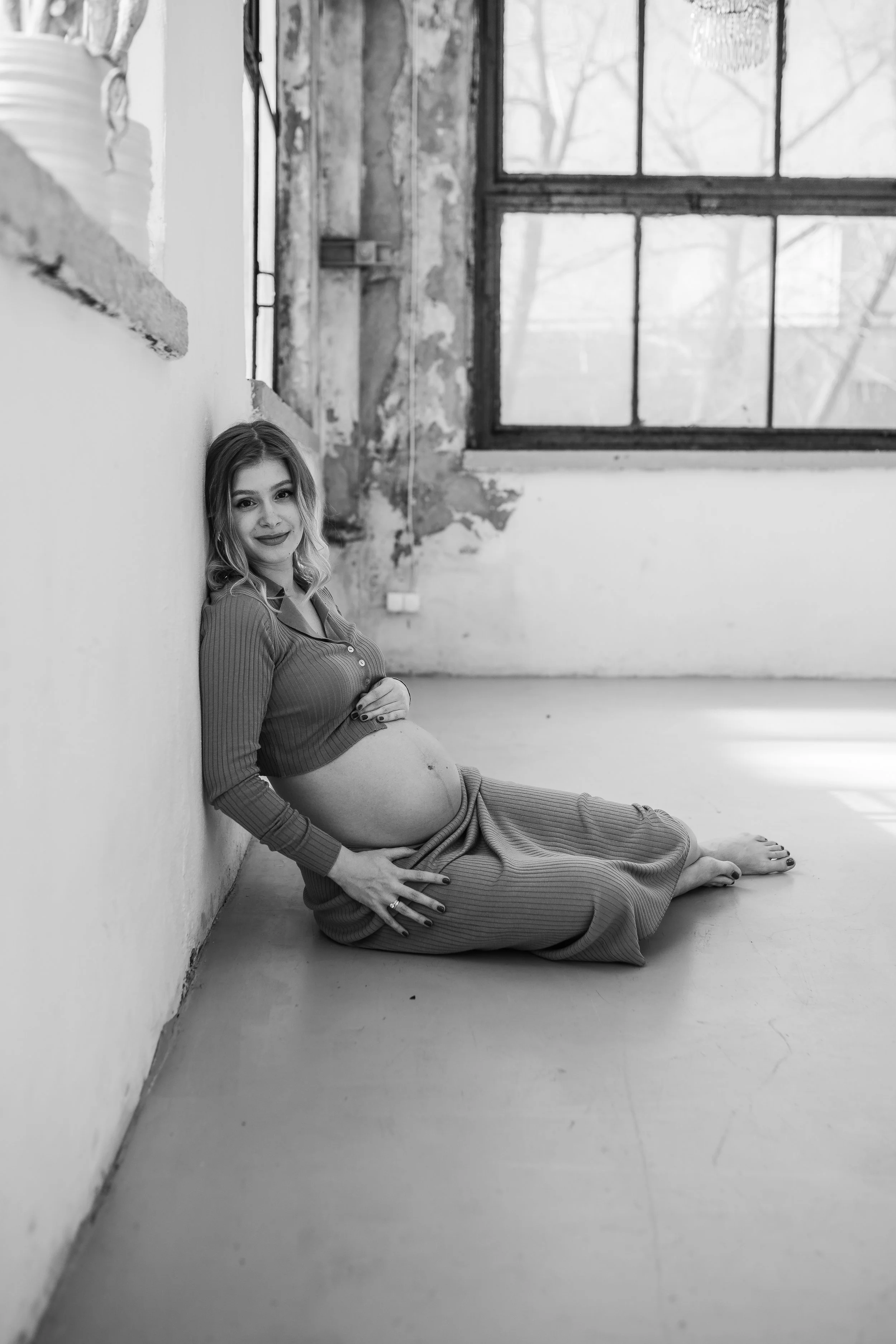 A pregnant woman with wavy hair, wearing a long-sleeved top and a long skirt, sitting on the floor, resting against a wall in a room with large windows and an unfinished wall behind her.