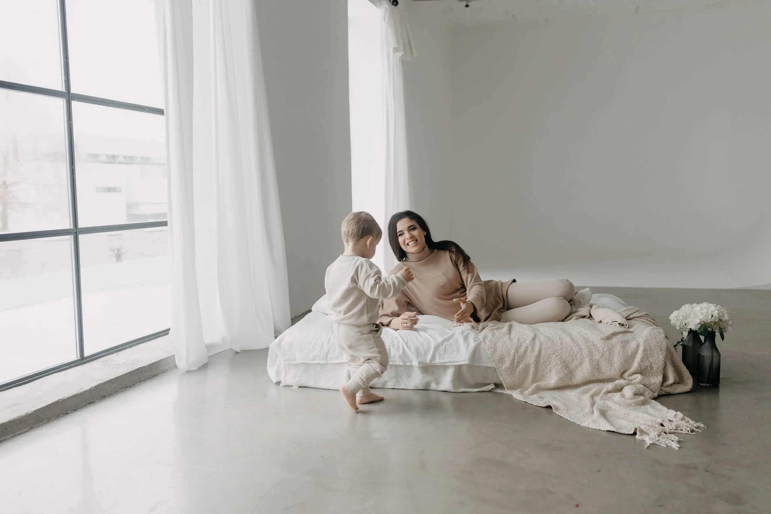 A woman and a young child playing on a bed in a bright, minimalistic room with large windows and white curtains.