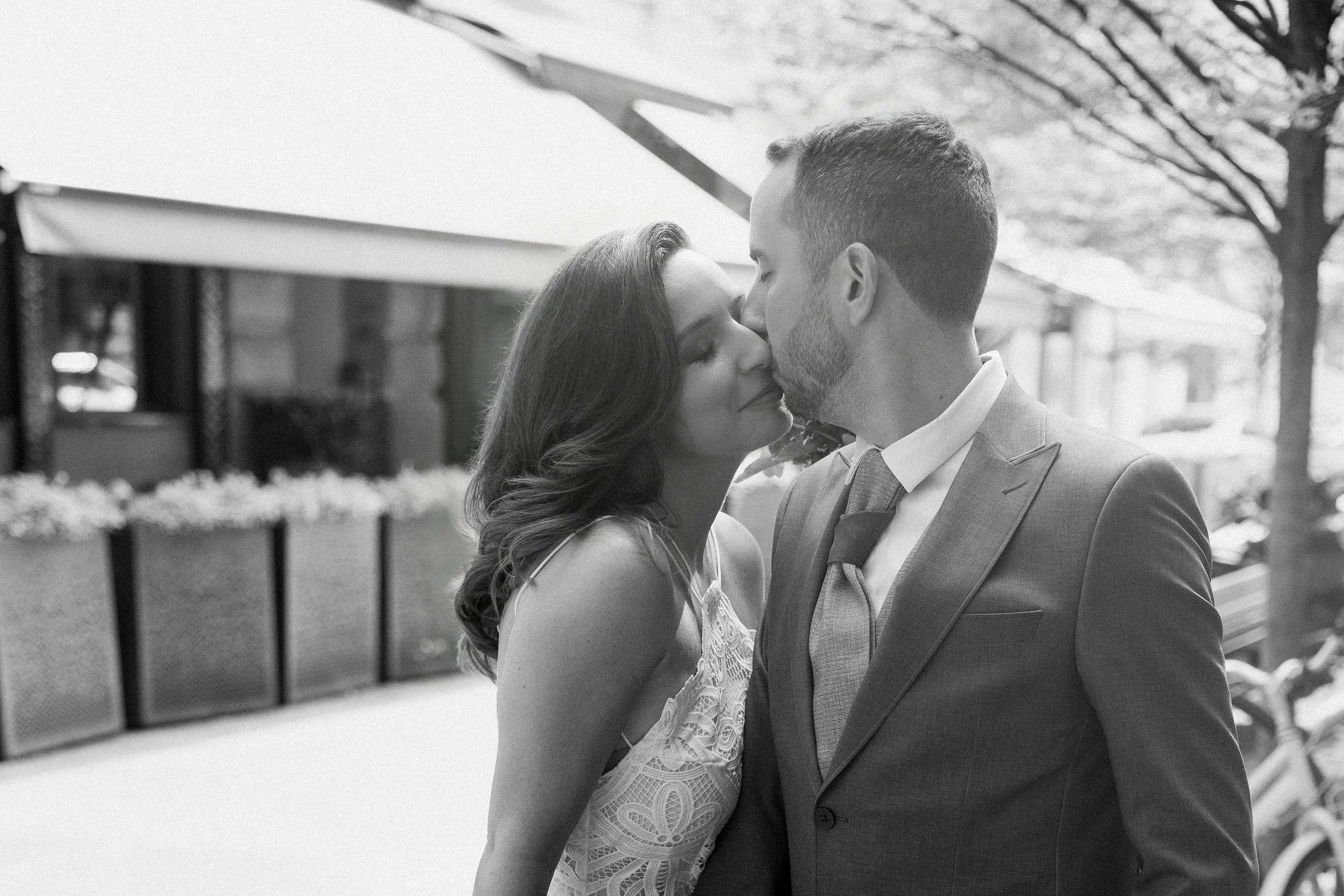 A black and white photo of a couple sharing a kiss outdoors, with the woman in a lace dress and the man in a suit, standing near a railing and trees.