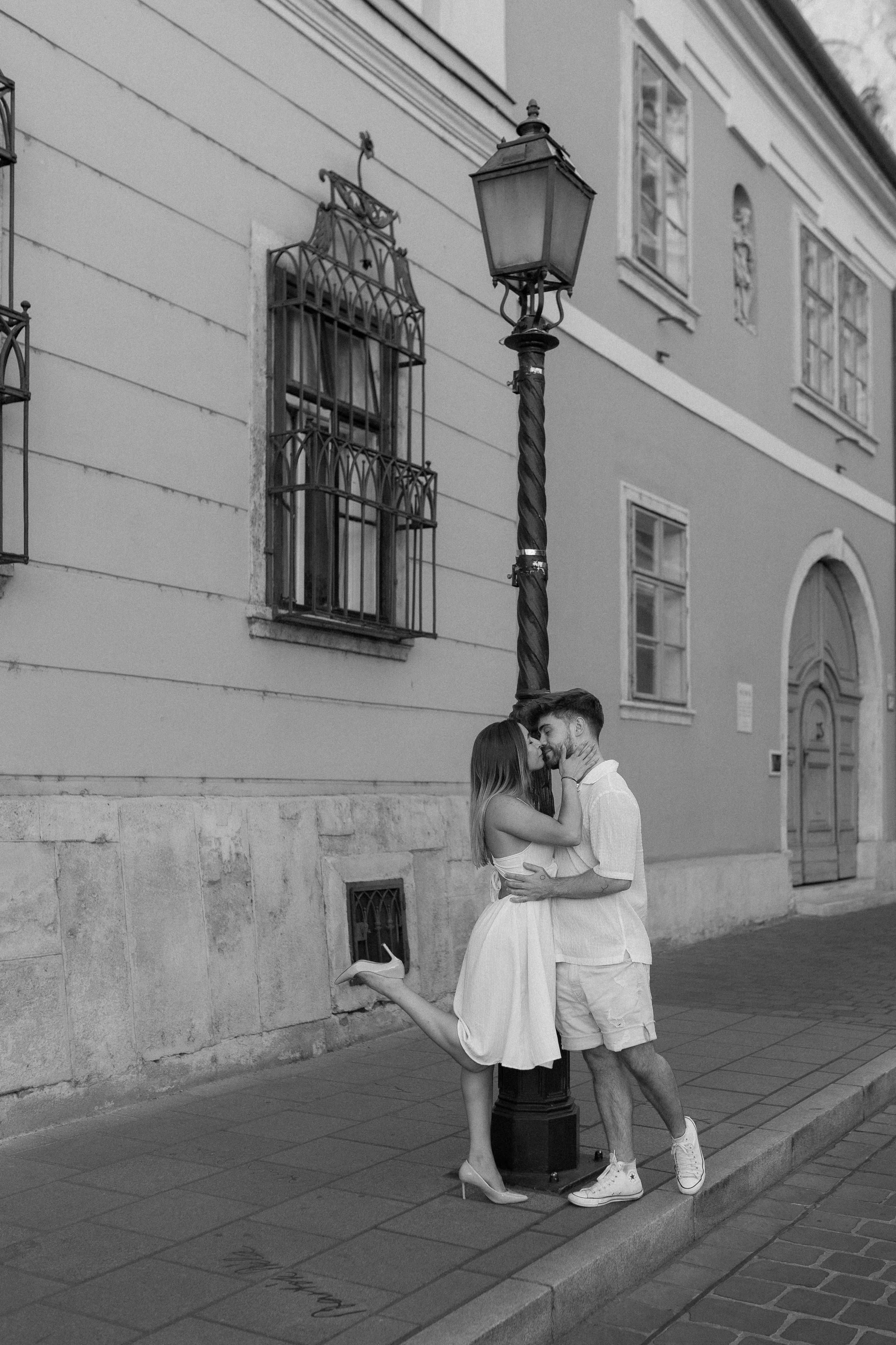 A couple dances on a city sidewalk at night, embracing each other near a lamp post, with old buildings and stone wall behind them.