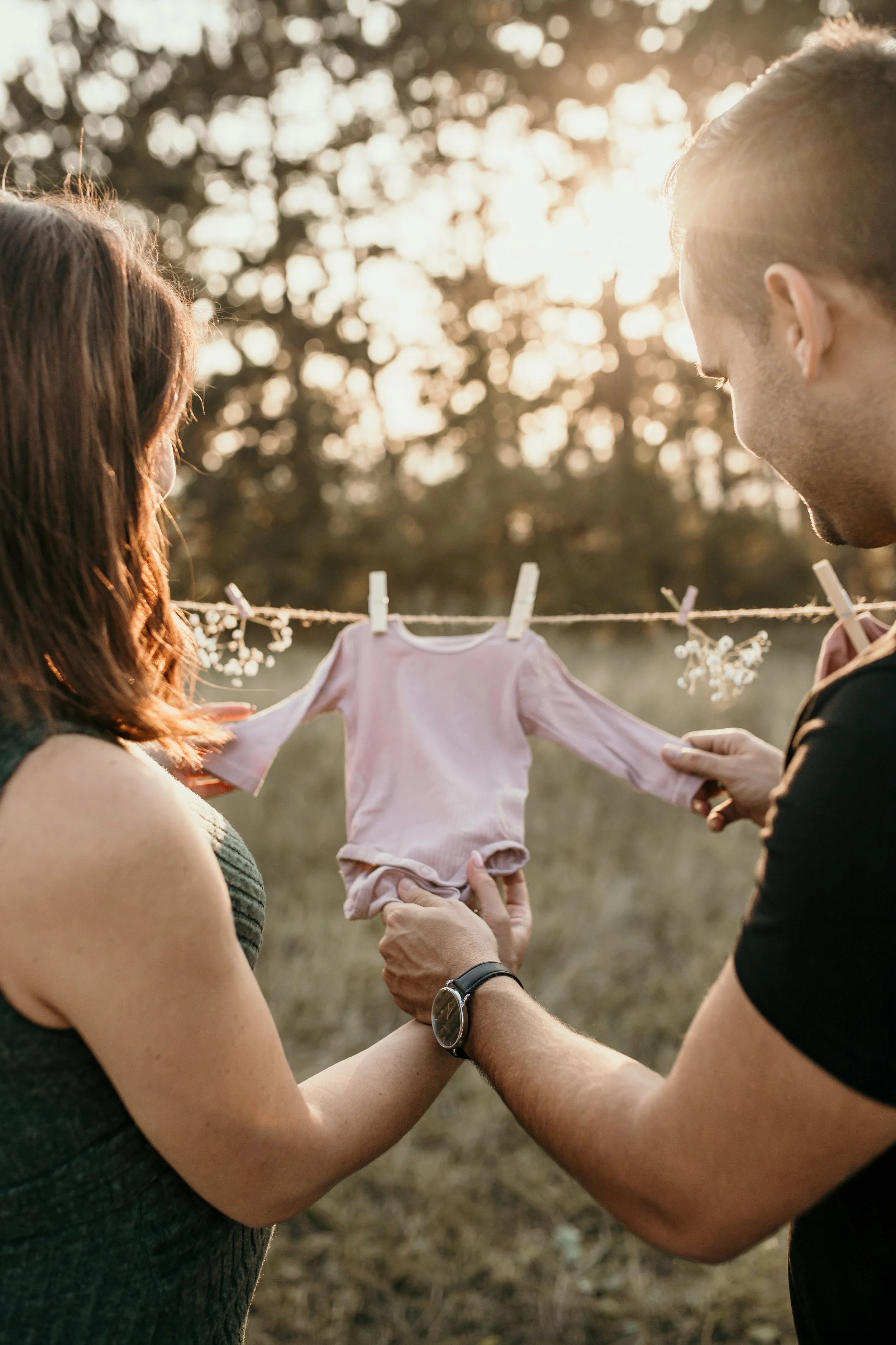 A couple holding a baby outdoors during sunset, with the baby hanging between them on a clothesline decorated with small flowers.
