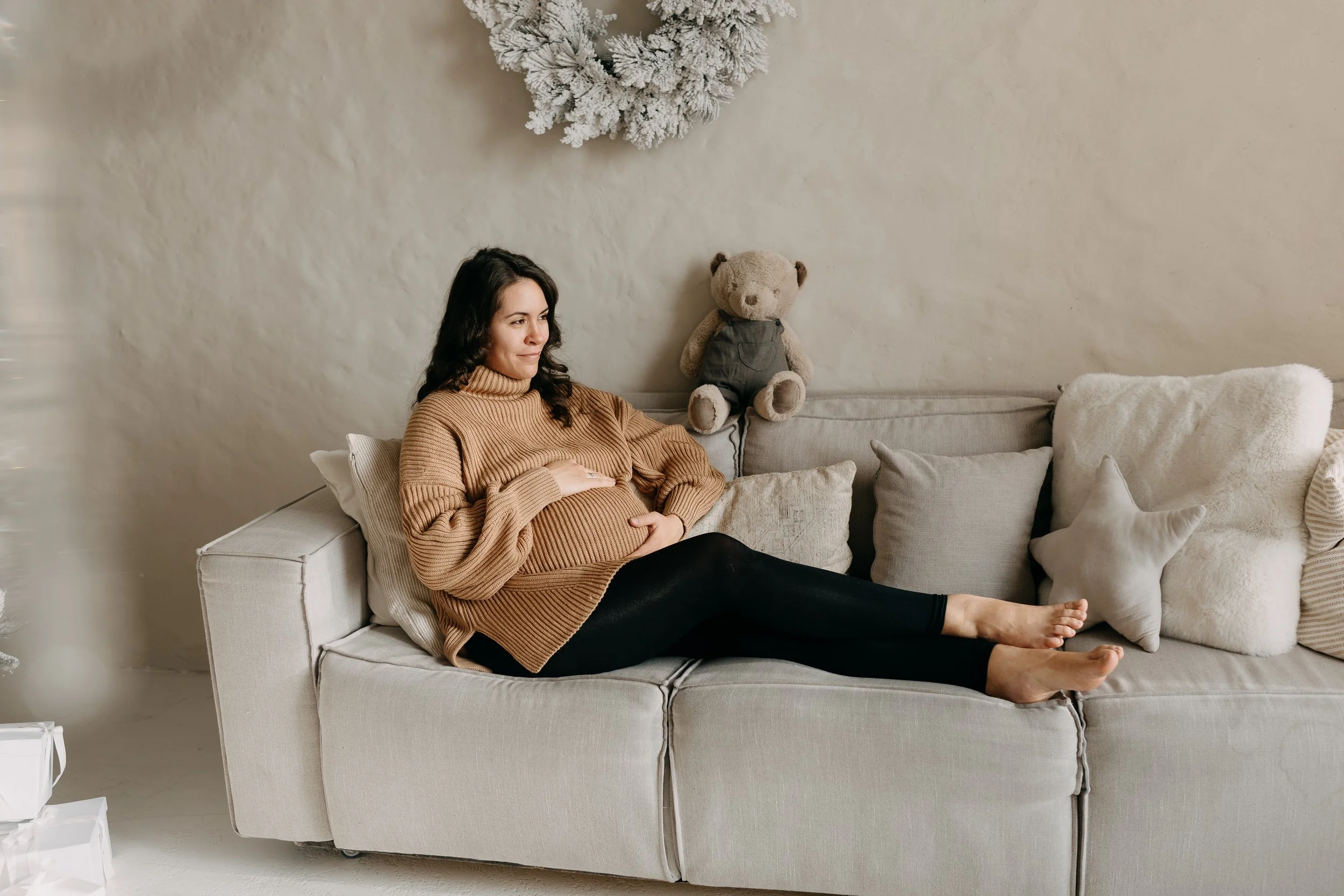 A pregnant woman sitting on a light-colored couch, with a stuffed bear and pillows behind her, in a room decorated with a white, snow-covered wreath on the wall.