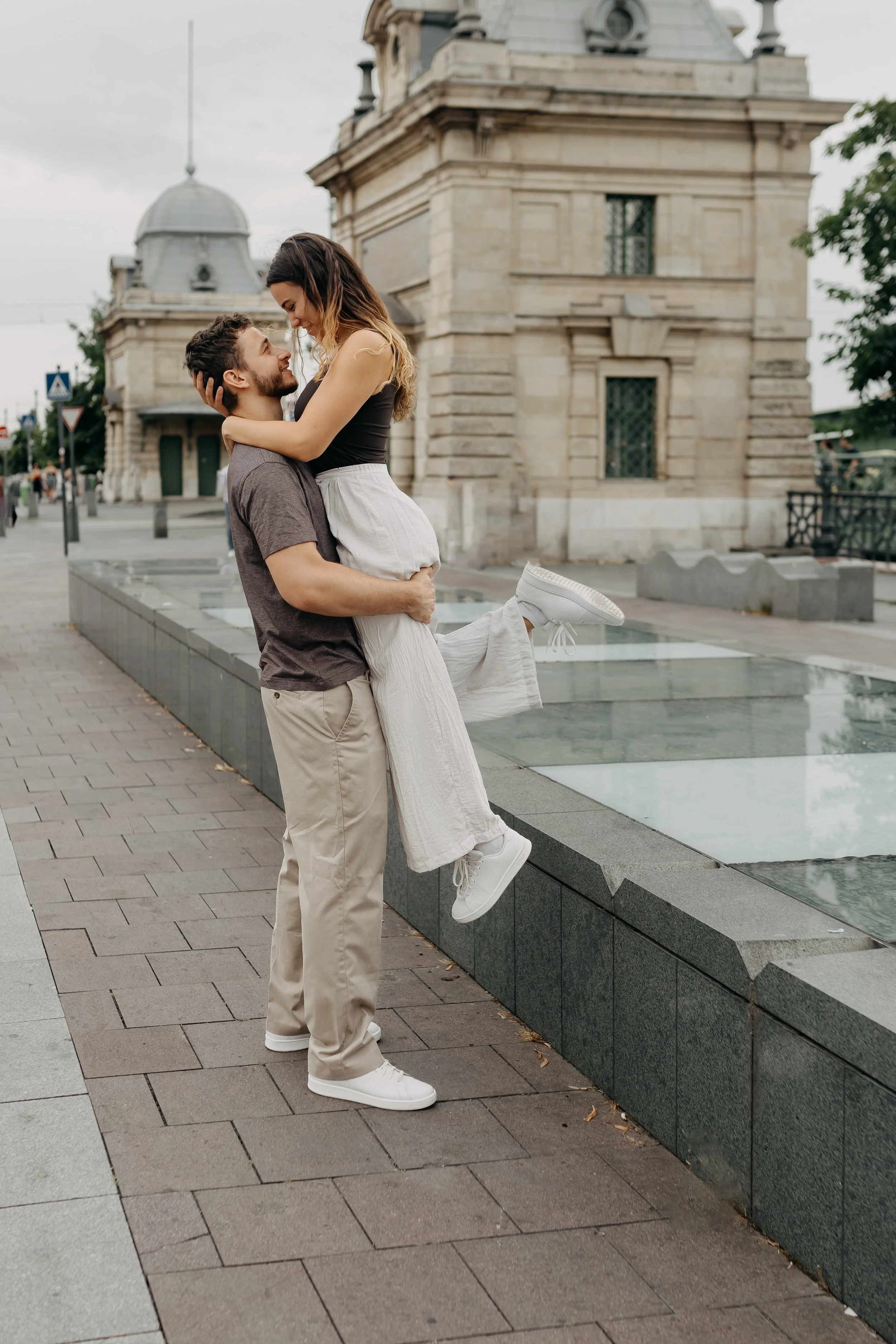 A happy couple with the man holding the woman who is smiling and looking down at him, in an urban area with historic buildings and paved walkways, overcast sky.