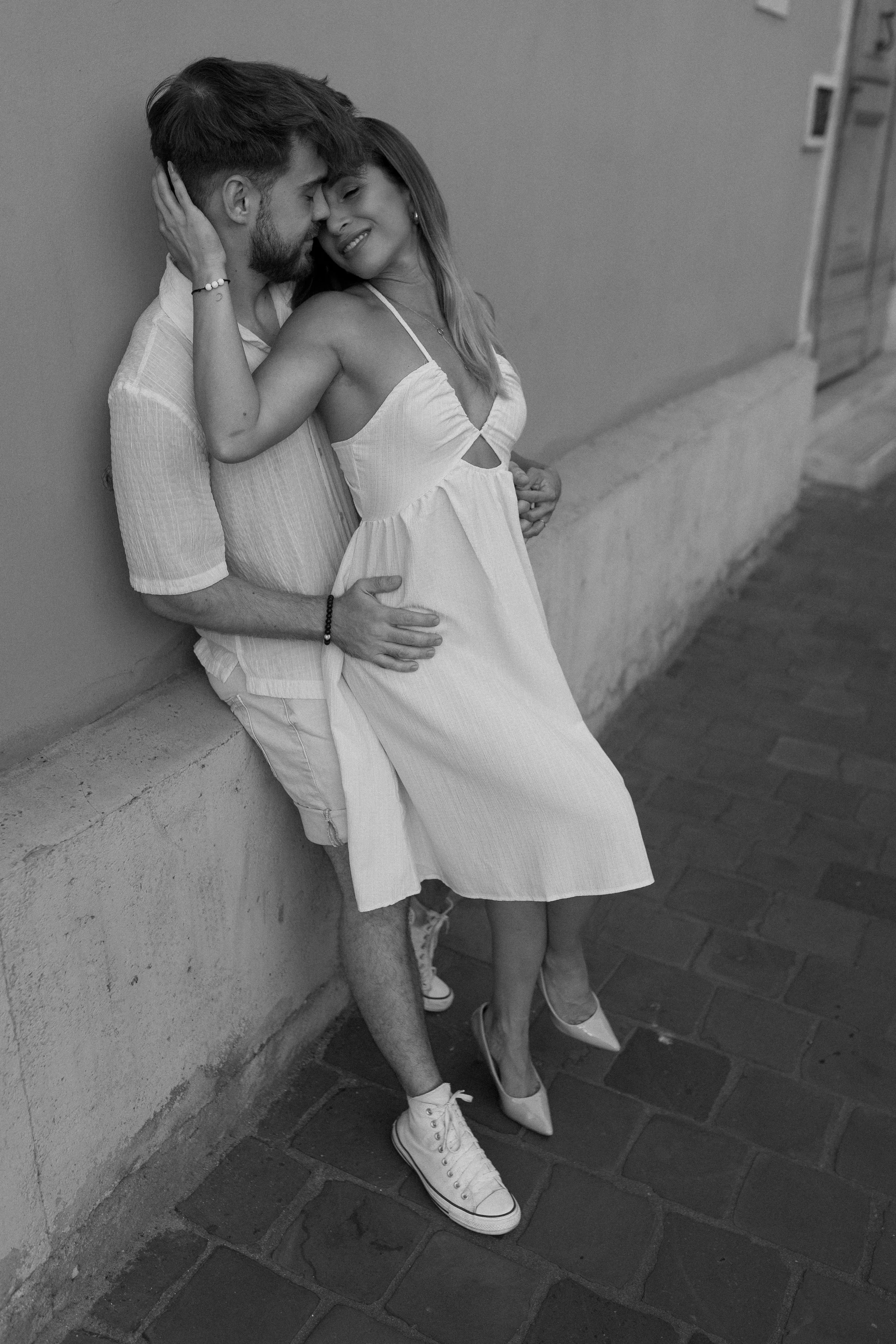 A black and white photo of a man and woman leaning against a wall. The man is wearing shorts and sneakers, while the woman is in a dress and heels. They are close, with the woman smiling and the man gently touching her face.