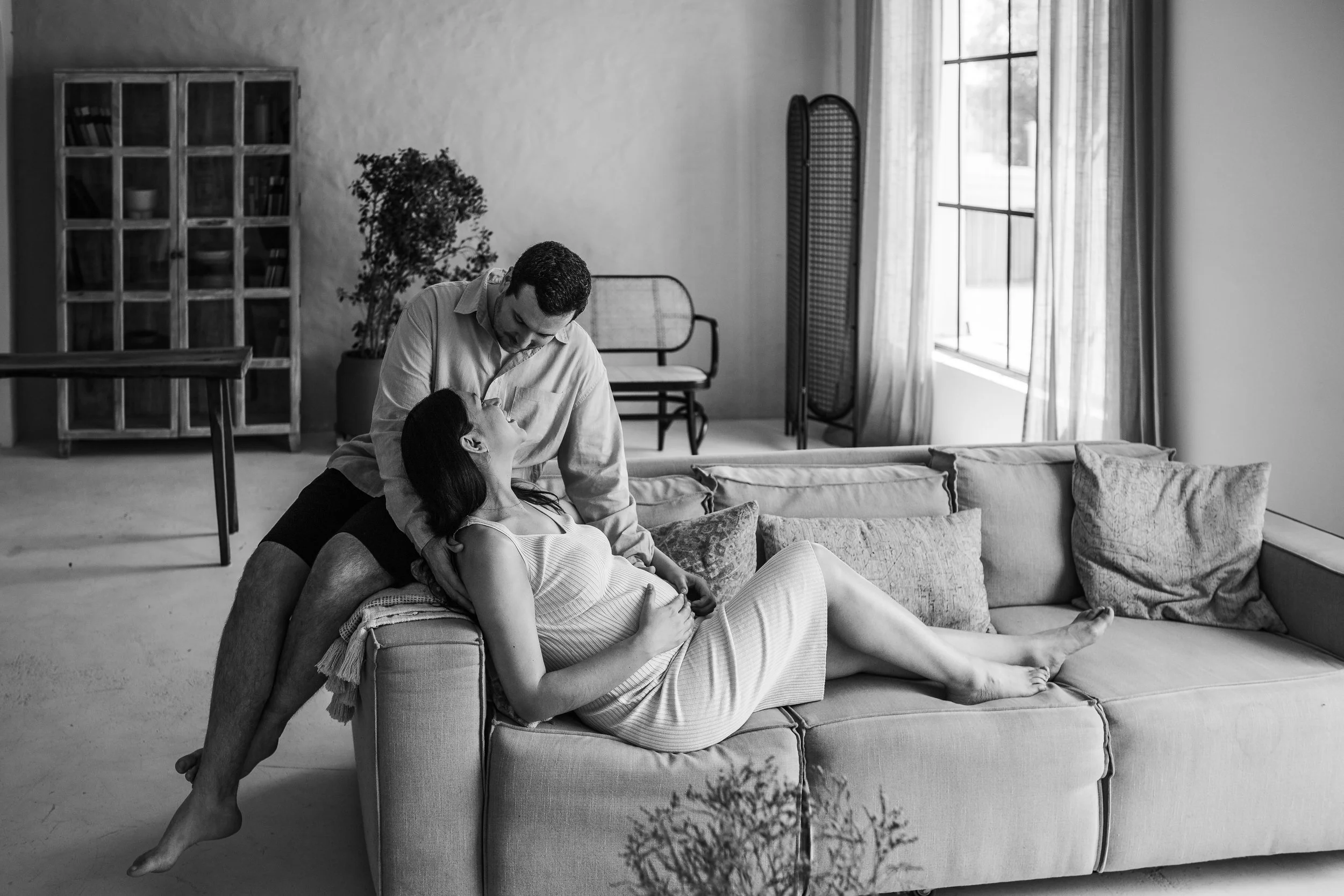 A couple engaging intimately on a sofa in a well-lit living room, with large windows and a folding screen in the background.