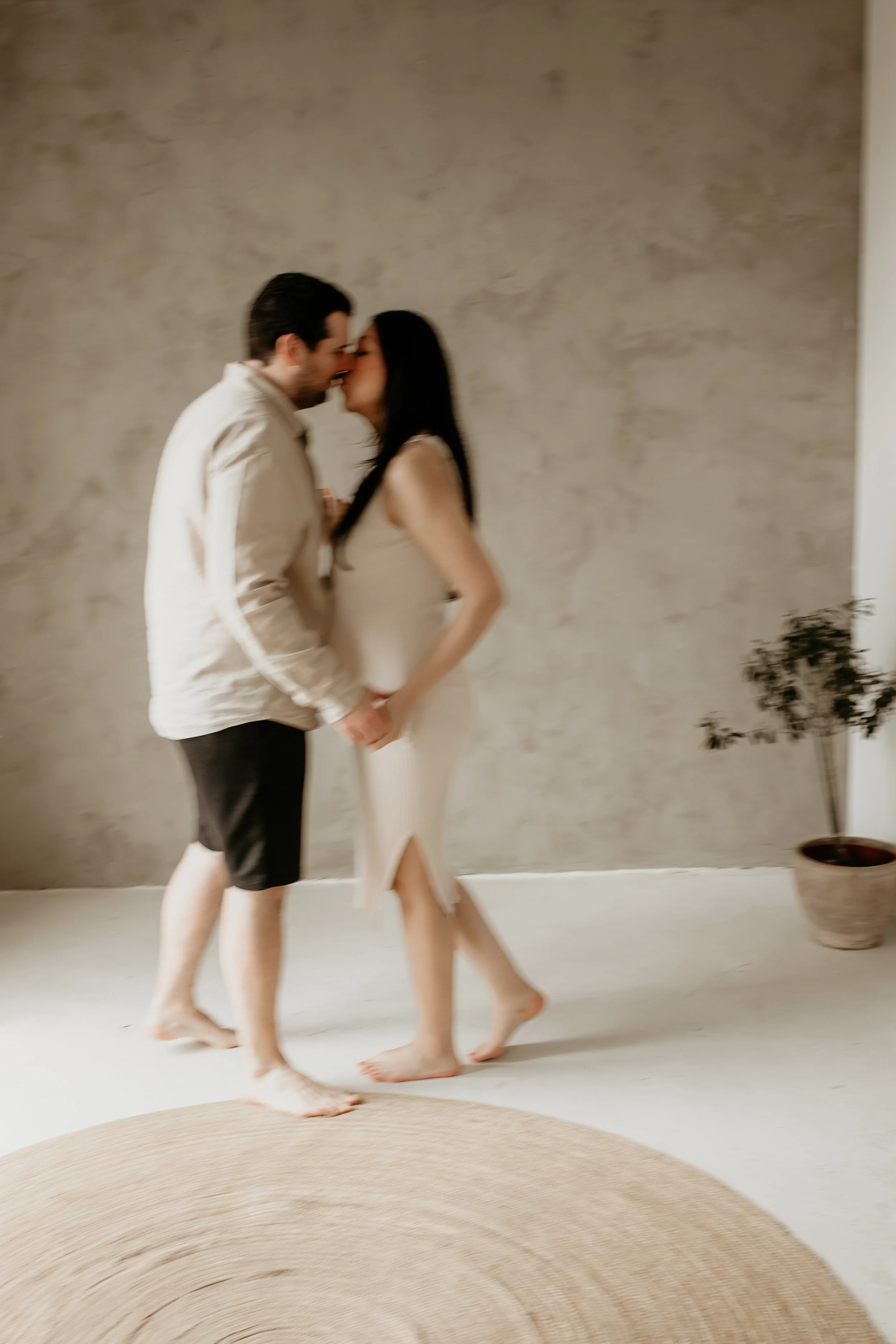A couple is dancing closely indoors, holding hands, with a beige rug in the foreground and a potted plant in the background.