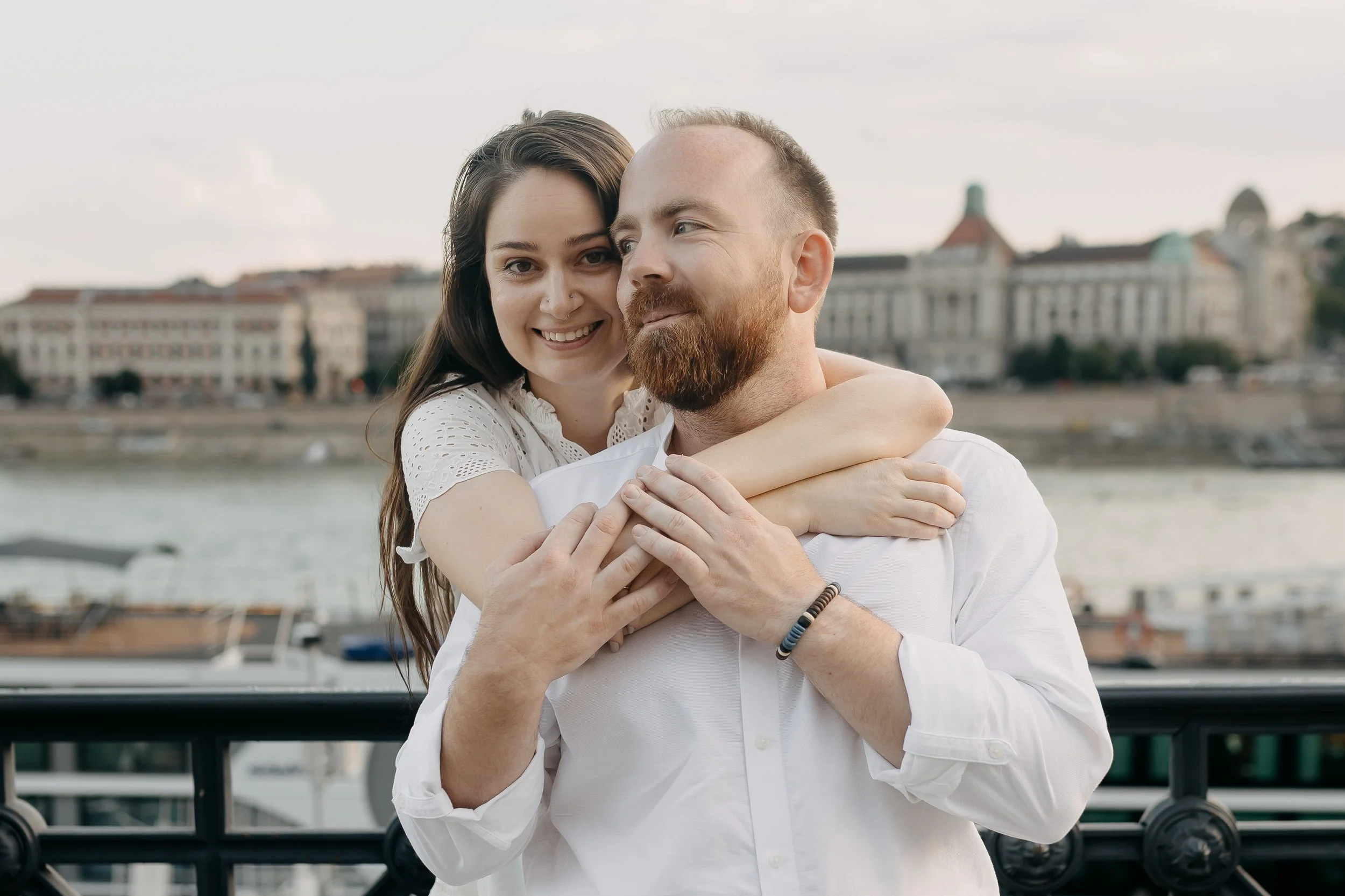 A couple hugging each other outdoors with water and city buildings in the background during daytime.