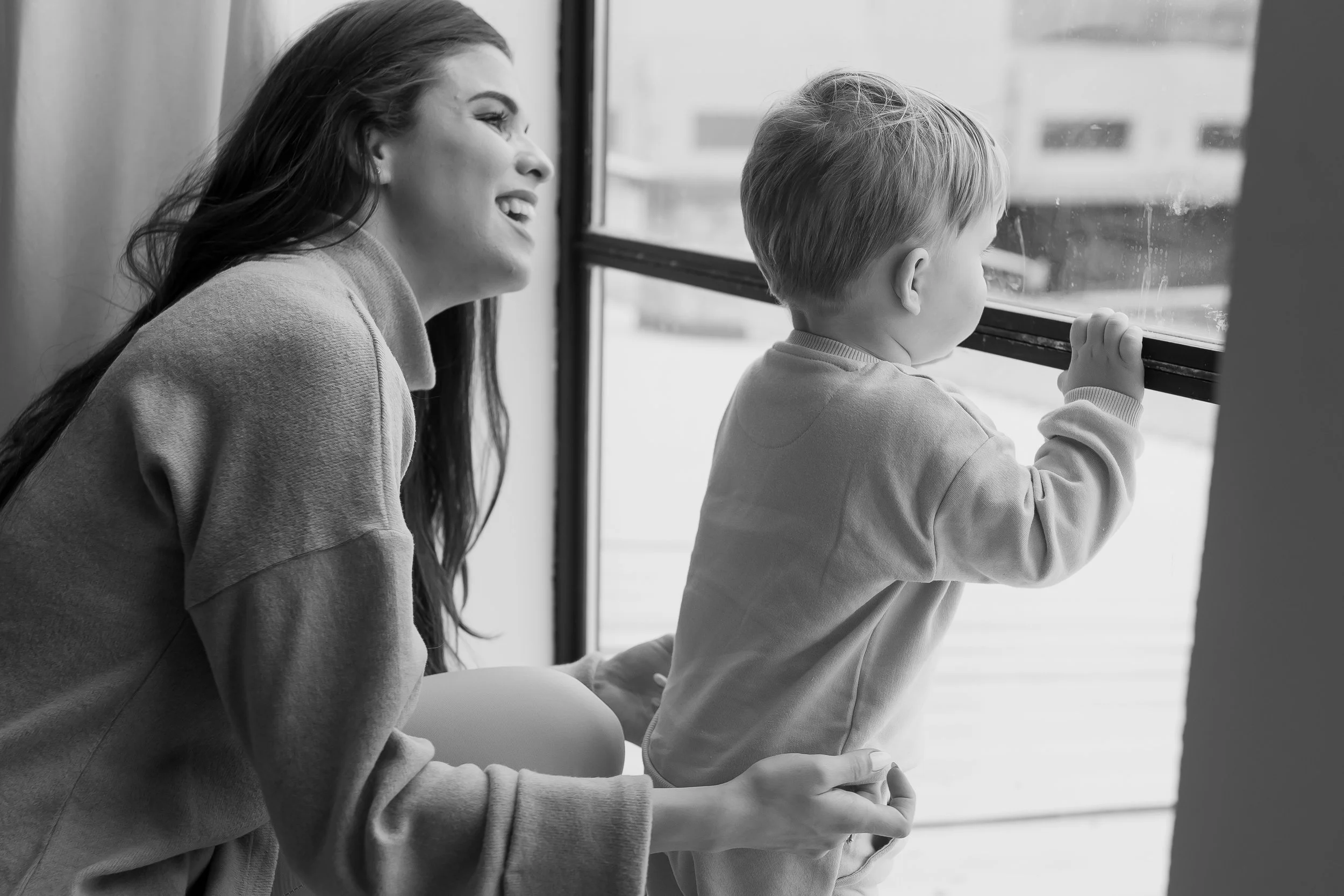 A woman smiling and sitting beside a young boy who is looking out a window, holding the window frame with one hand, in a black and white photo.