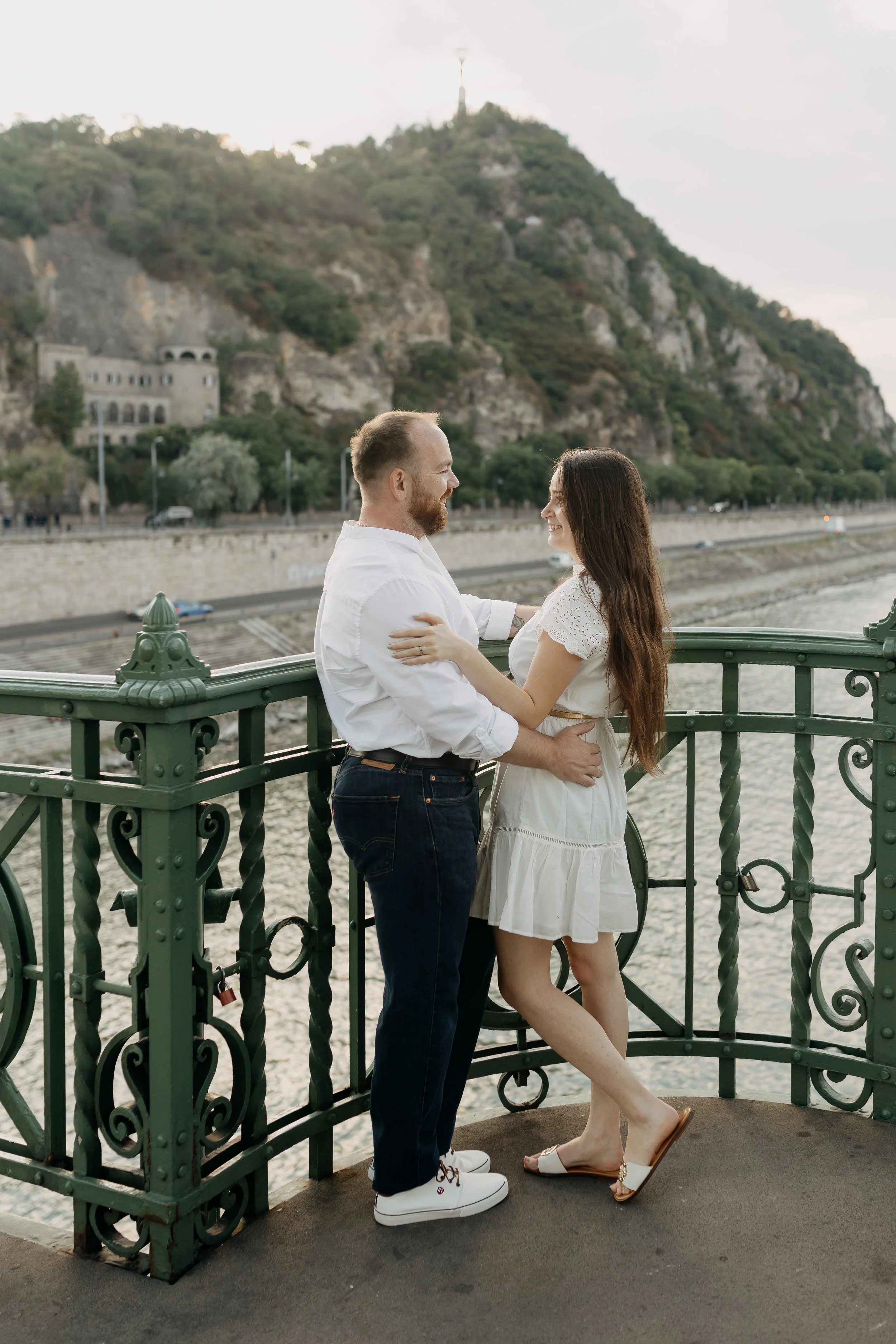 A couple standing close together on a bridge, smiling at each other, with a scenic background of a river, trees, and a hill with a statue on top.