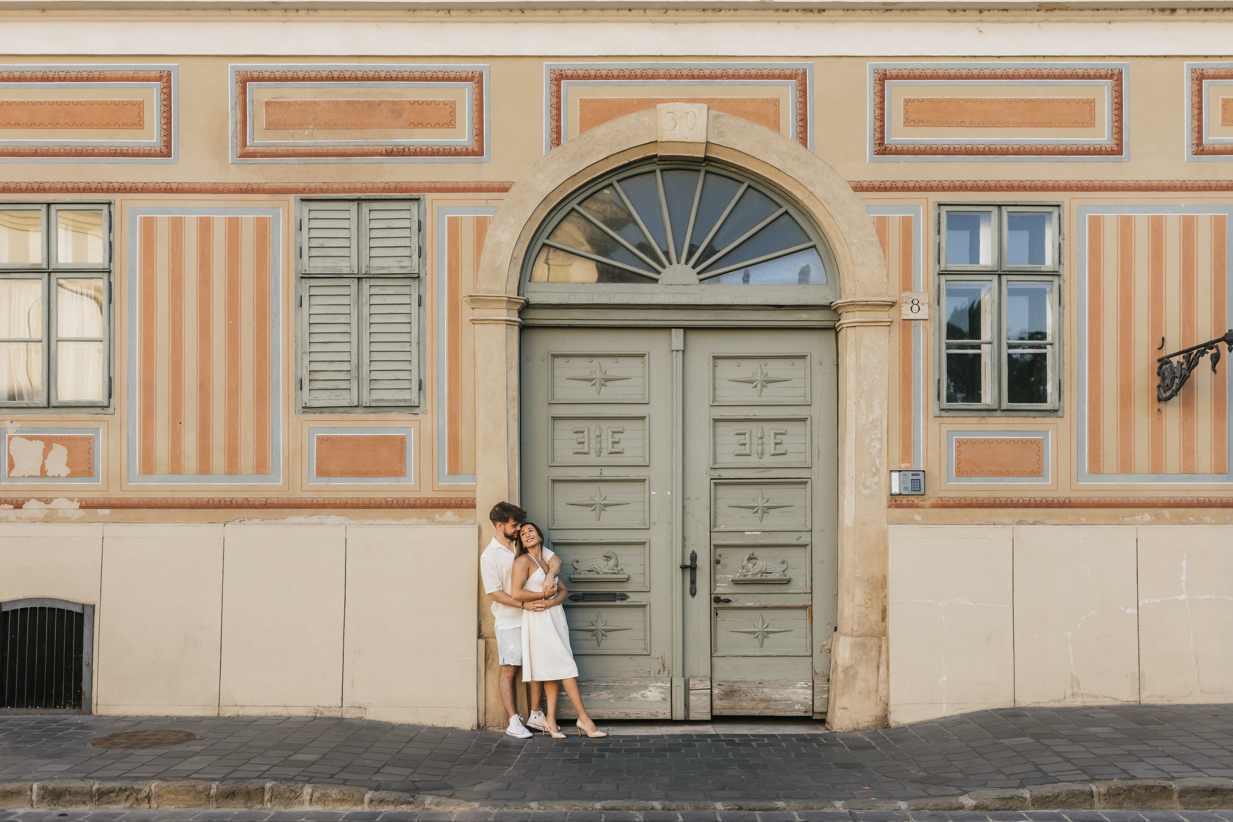 A couple stands in front of an ornate, large green door with a decorative fan-shaped window and intricate trim, against a peach-colored building with striped wall panels and shutters. The man, dressed in white, gently holds the woman, who wears a whi
