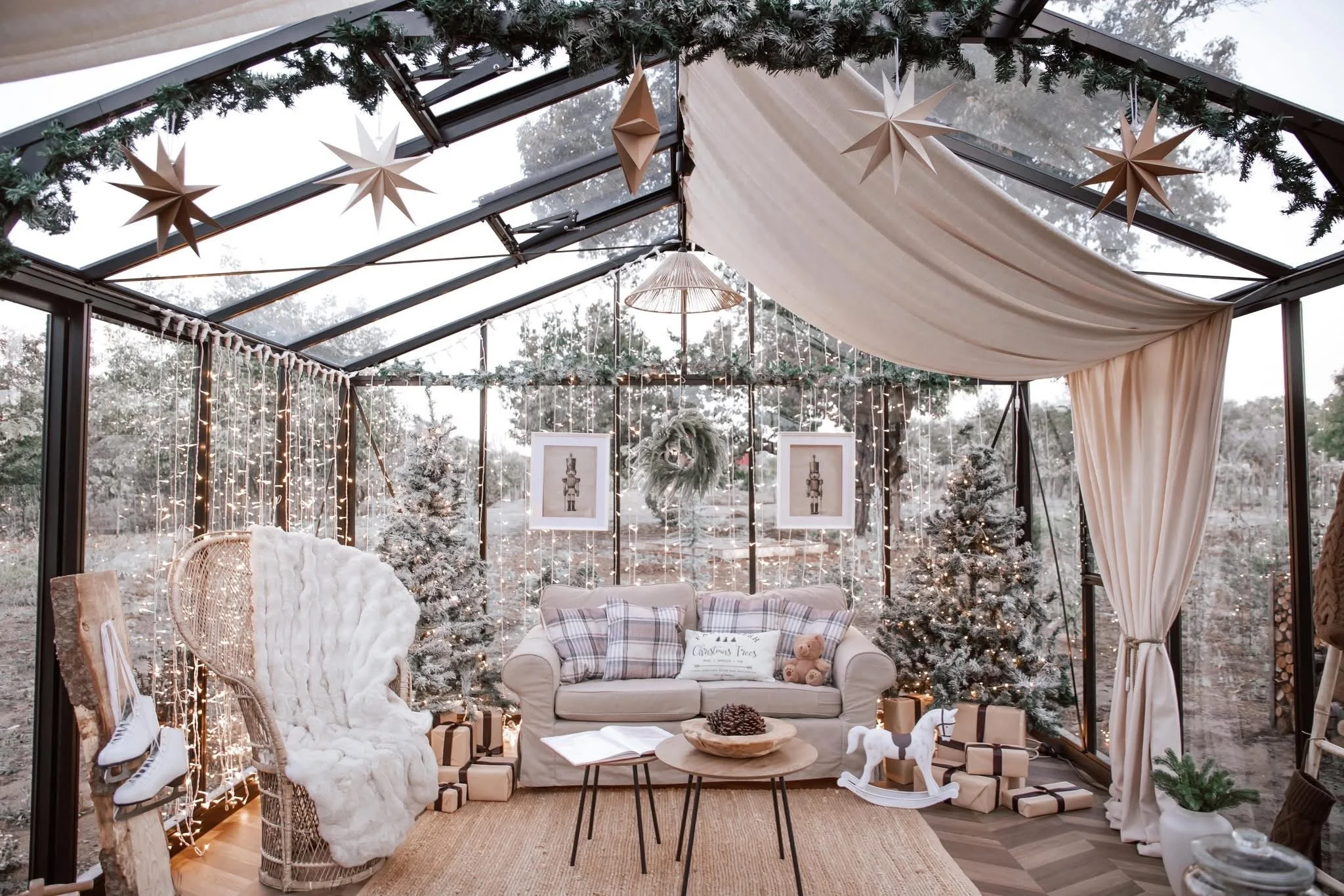A cozy, decorated indoor living space with Christmas and winter decorations, including fairy lights, snow-covered trees, and hanging stars. There are curtains and framed pictures on the back wall, and furniture like a sofa, armchair, and a small table with pinecones and a wooden horse.
