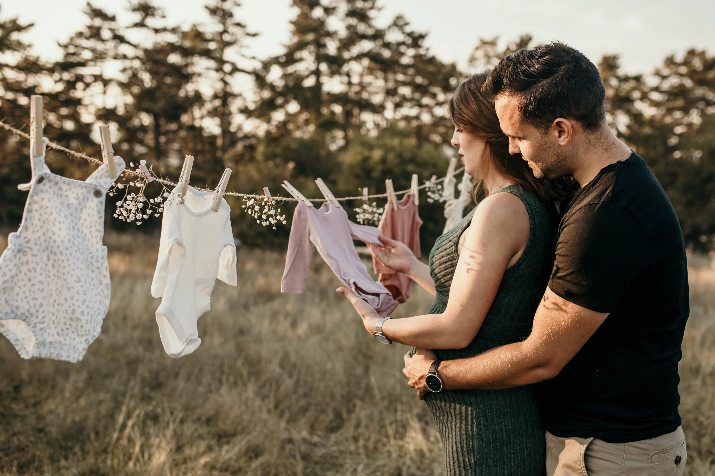 A couple standing outdoors by a clothesline with baby clothes hanging, with trees in the background.