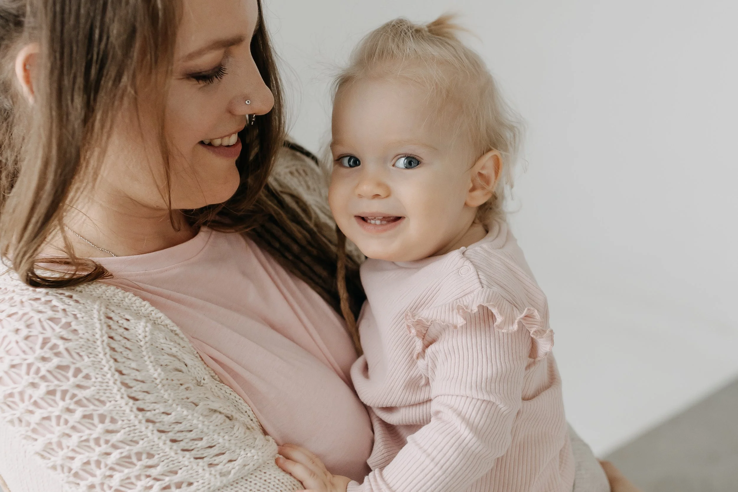 A woman with long brown hair, a nose piercing, and wearing a pink shirt with a beige crochet cardigan holding a young girl with curly blonde hair, blue eyes, and wearing a pink shirt with ruffle details, smiling and looking at the camera.