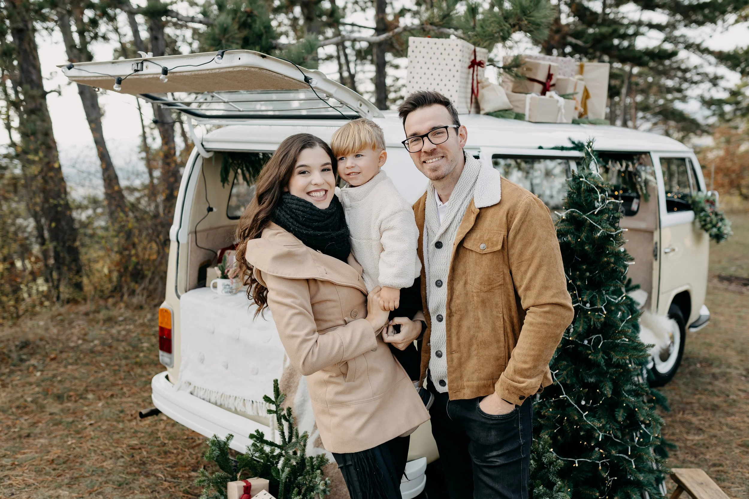 A smiling family of three standing in front of a vintage van decorated for Christmas with presents, a Christmas tree, and festive lights outdoors among trees.