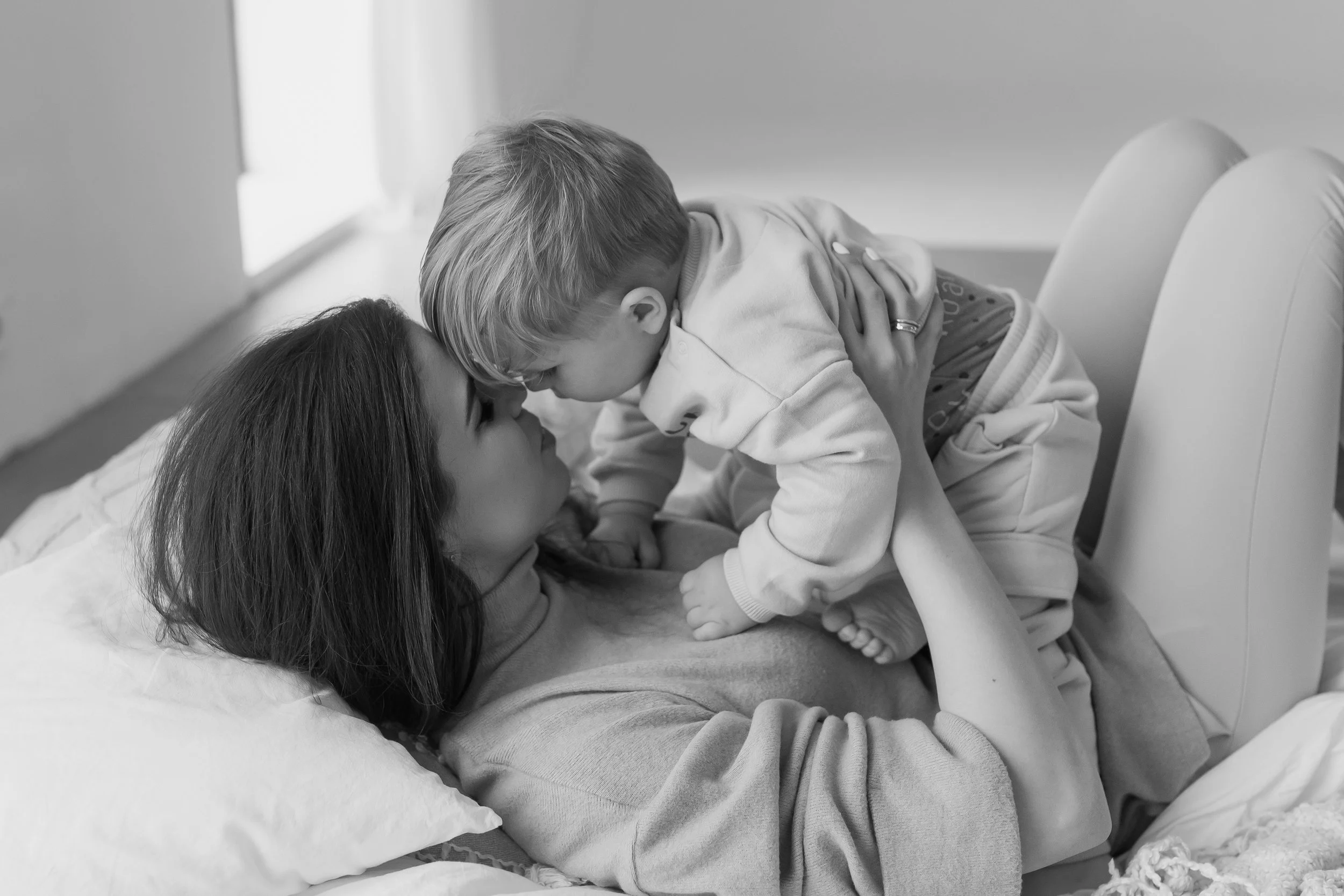 A woman lying on a hospital bed with a young boy, presumably her son, leaning over her to give a kiss on her forehead. The woman is resting her head on a pillow, and the boy is on her chest, both showing an emotional connection.