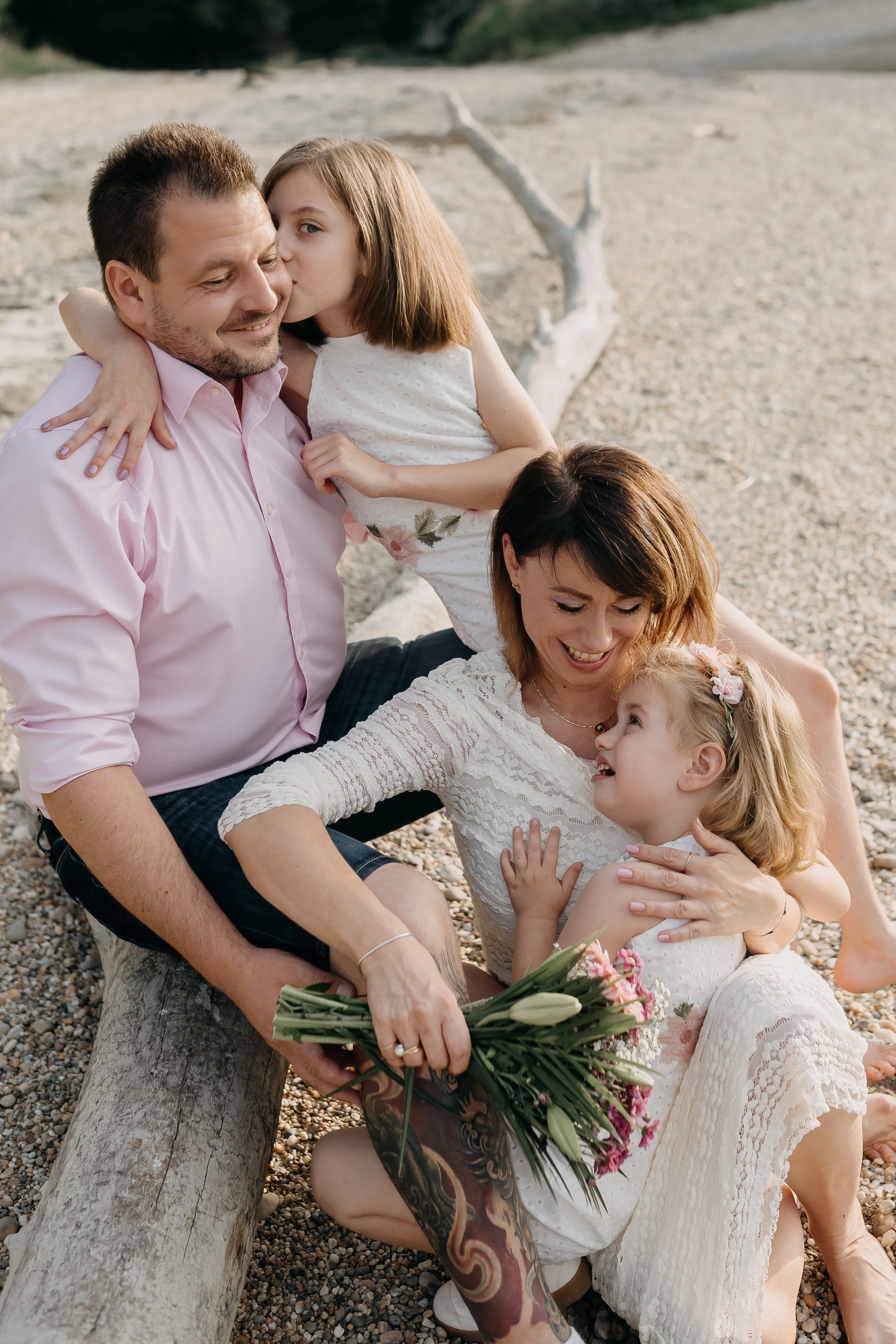 A family of four enjoying a moment on a beach with a driftwood log, with a man, woman, and two young girls, one of whom is giving a kiss to the man. The woman and one girl are sitting close on the ground, and the man is holding a bouquet of flowers.