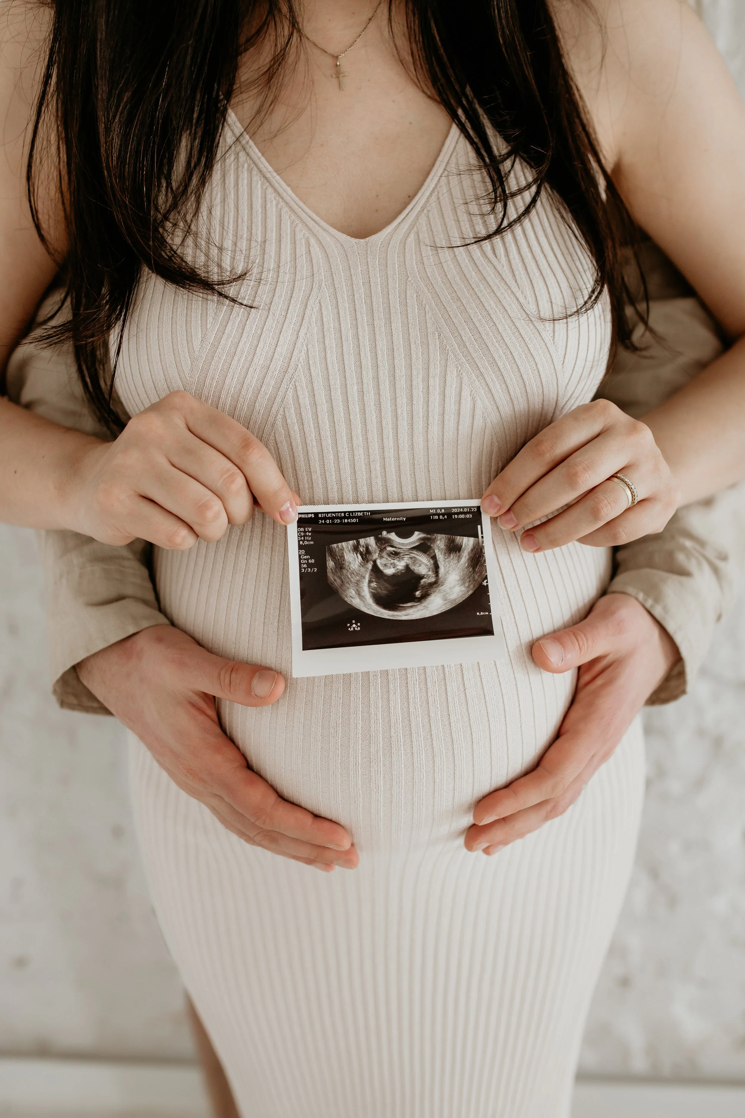 A pregnant woman in a beige ribbed dress holding an ultrasound image over her belly, with another person supporting her from behind.