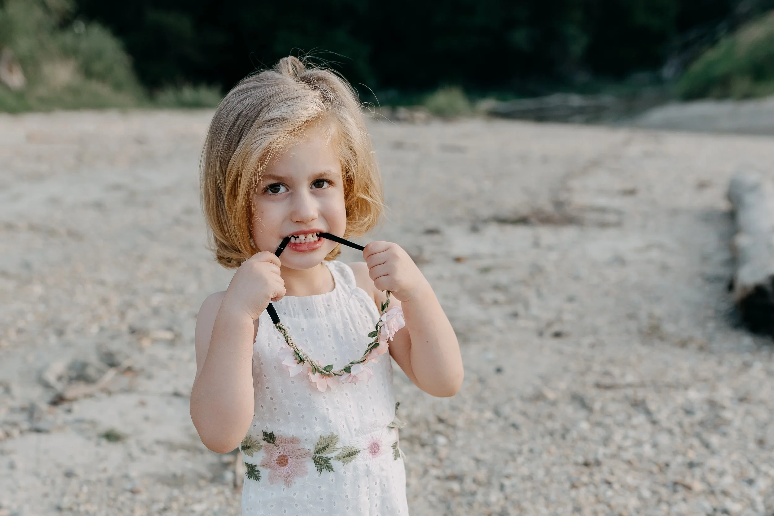 A young girl with blonde hair in a white dress holding sunglasses in her hands and standing on a beach with trees in the background.