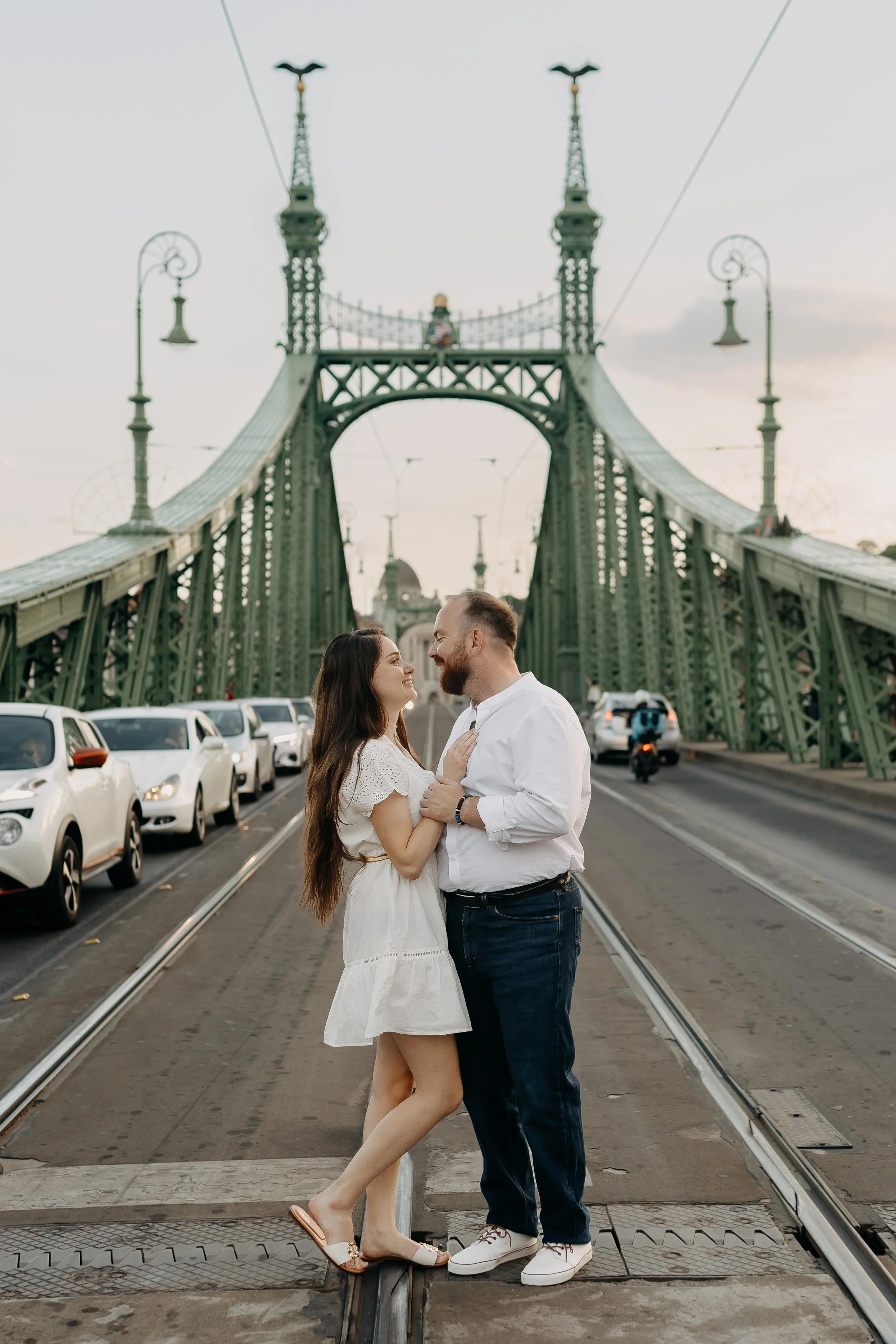 A couple standing close together on a pedestrian bridge in front of a historic suspension bridge, with cars parked along the street beneath. The woman has long brown hair and is wearing a white dress and sandals, while the man has a beard and is dres