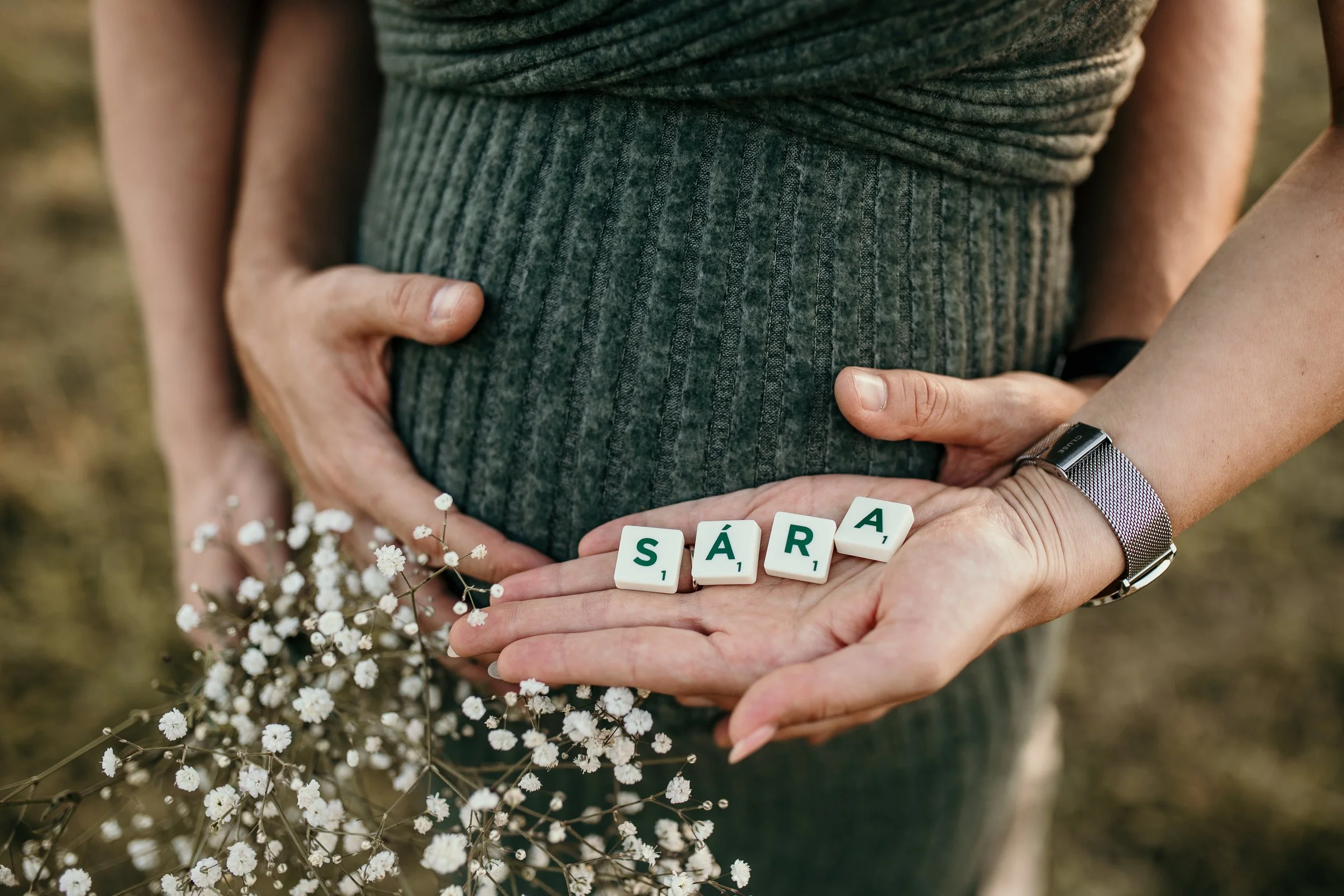Close-up of two people holding a pregnant woman's belly with one hand each, wearing gray clothing. There are six Scrabble tiles spelling 'SÁRA' on the hands, and some small white flowers nearby.
