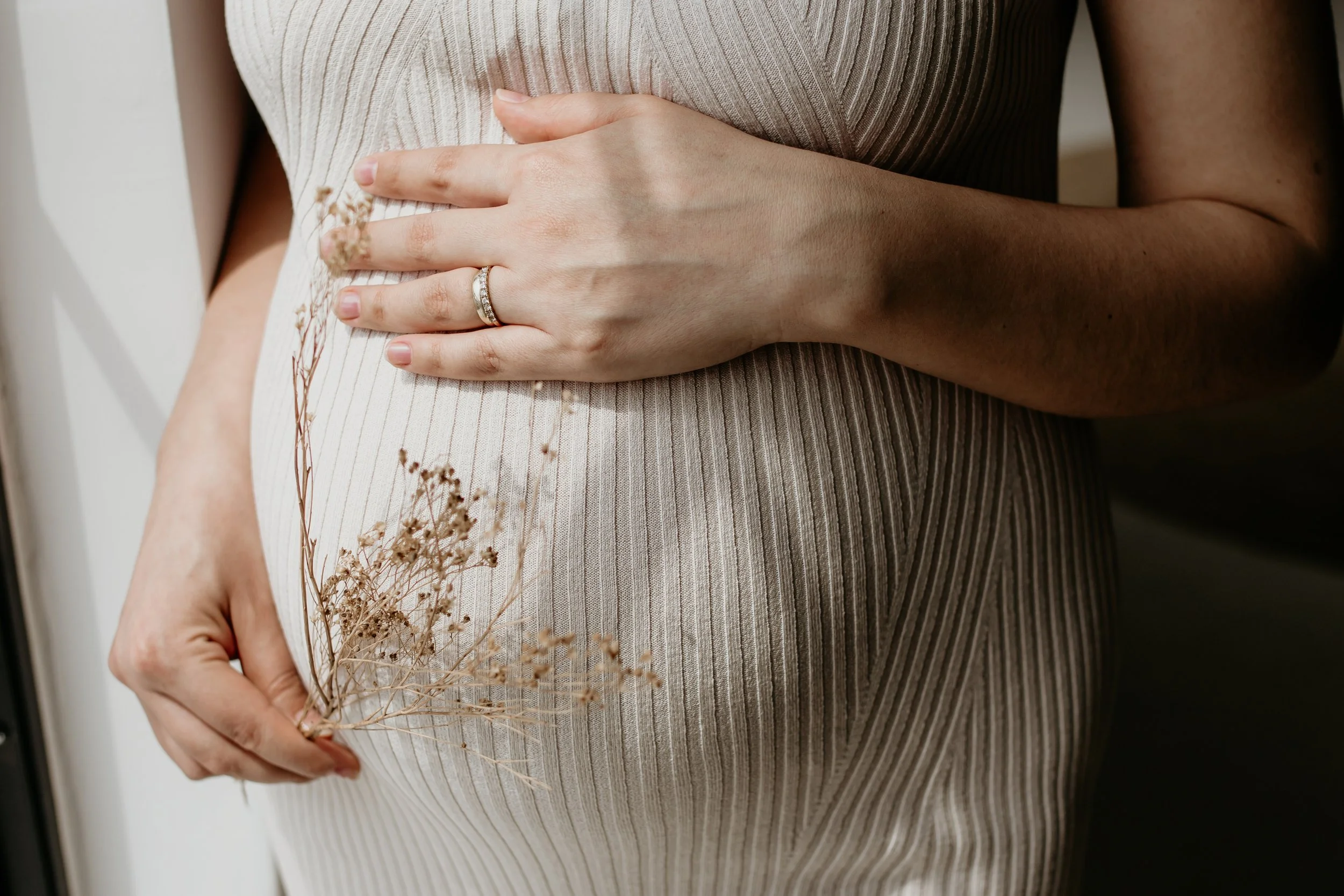Close-up of woman holding a small bouquet of dried flowers over her pregnant belly, wearing a beige ribbed dress, with her left hand displaying a wedding ring.