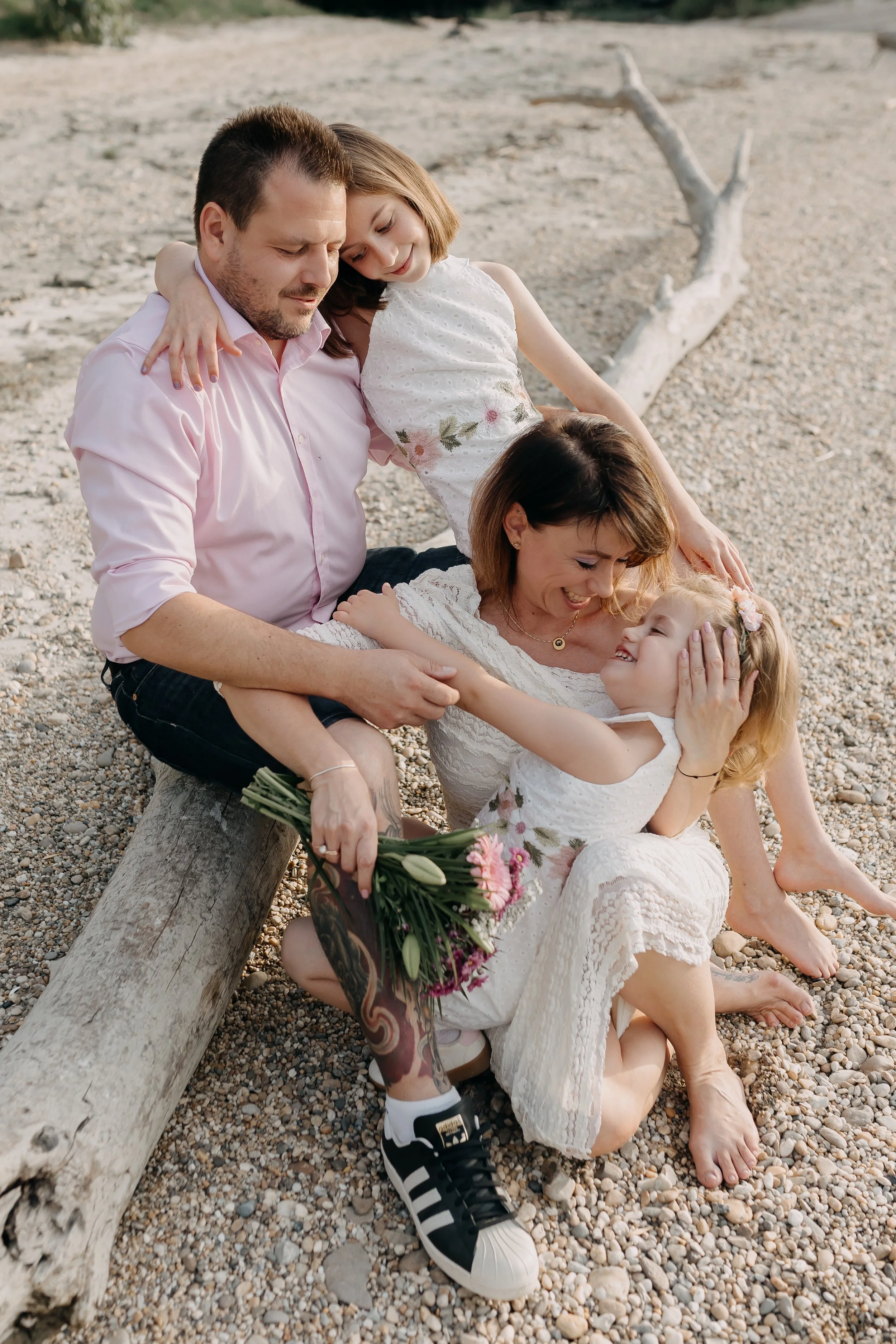 Four people, two women, one man, and a girl, sitting and lying on a pebble beach, smiling and engaging with each other. One woman is holding a bouquet of flowers. The scene is outdoors with a large piece of driftwood and some greenery in the backgrou
