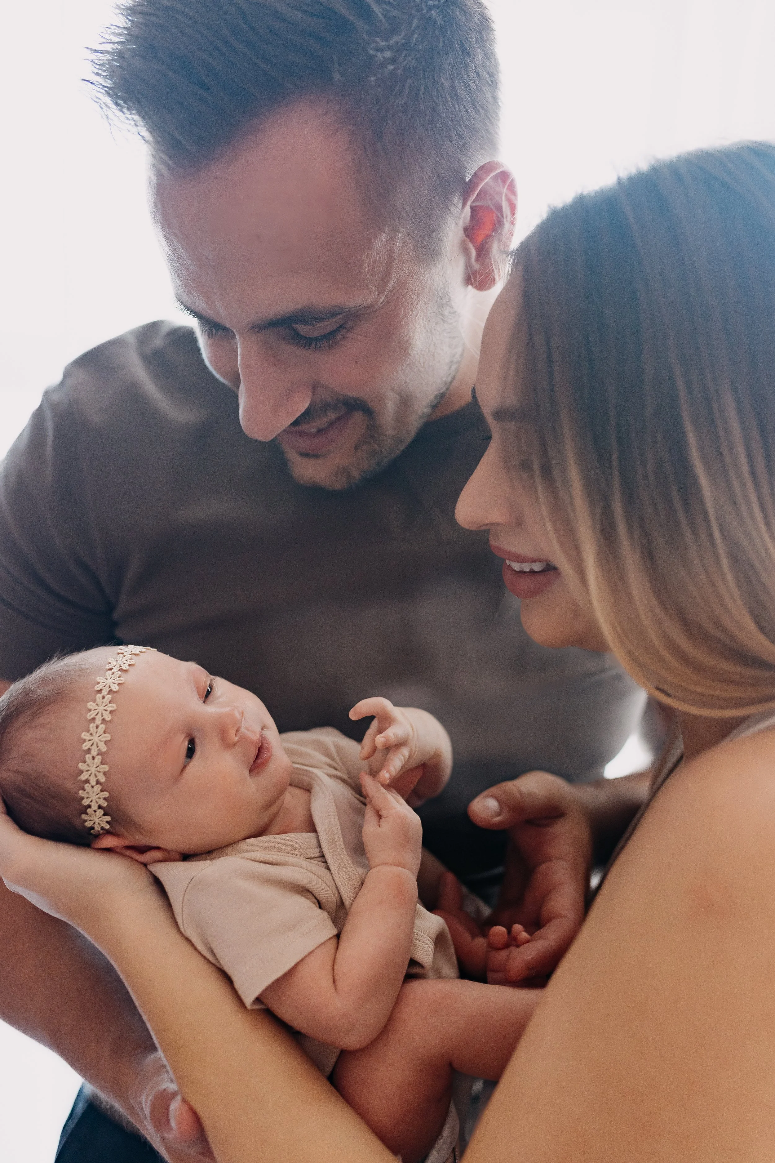 A couple holding and looking at a baby girl who is lying in their arms, wearing a beige outfit and a headband with flowers, indoors with soft natural light.