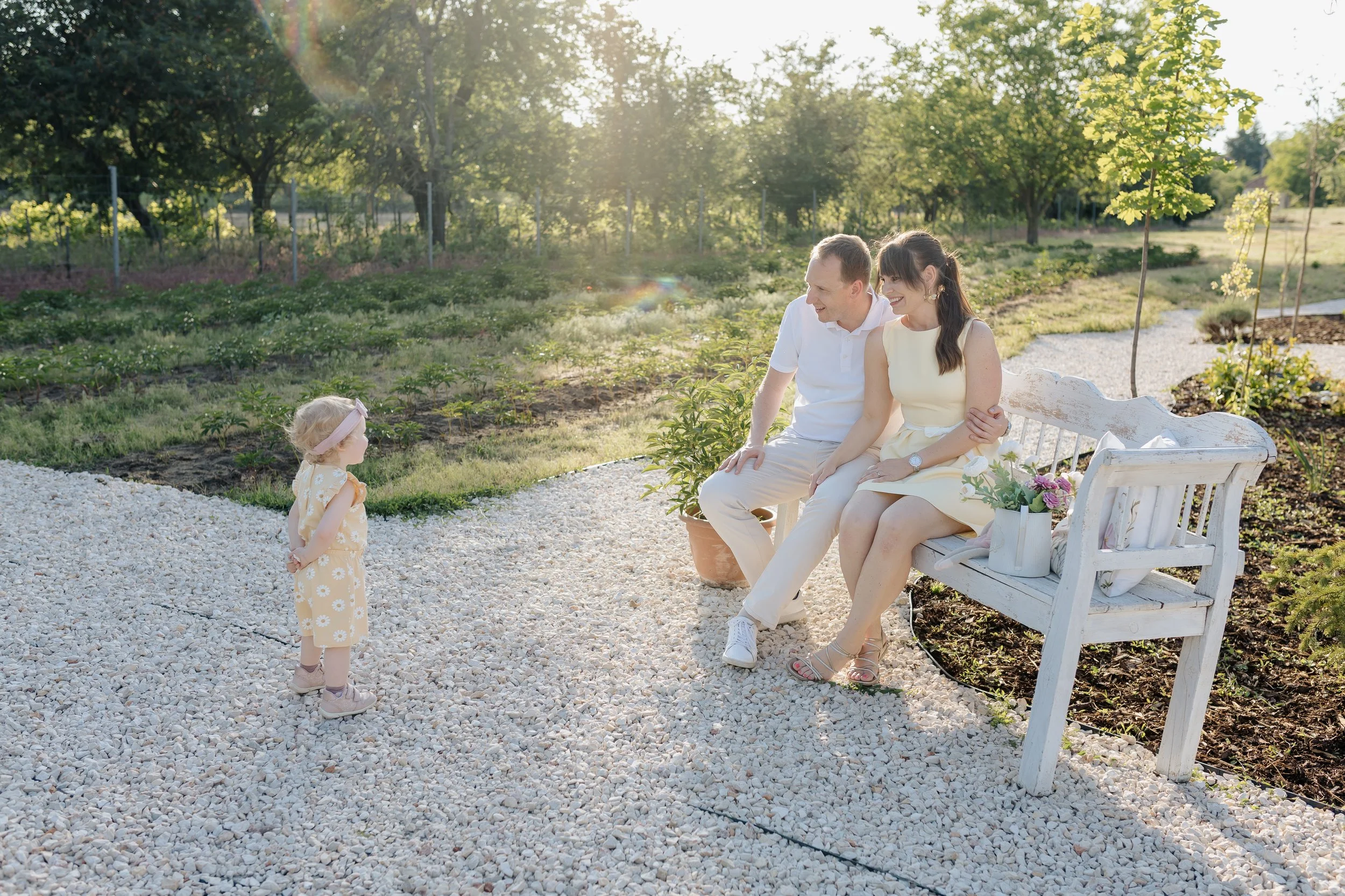 A family enjoying a sunny day in a garden. A man and woman are sitting on a white bench, smiling at a small girl who is standing nearby. The girl is wearing a yellow floral dress, and the woman is in a light yellow dress. There are trees and greenery in the background with sunlight streaming in.