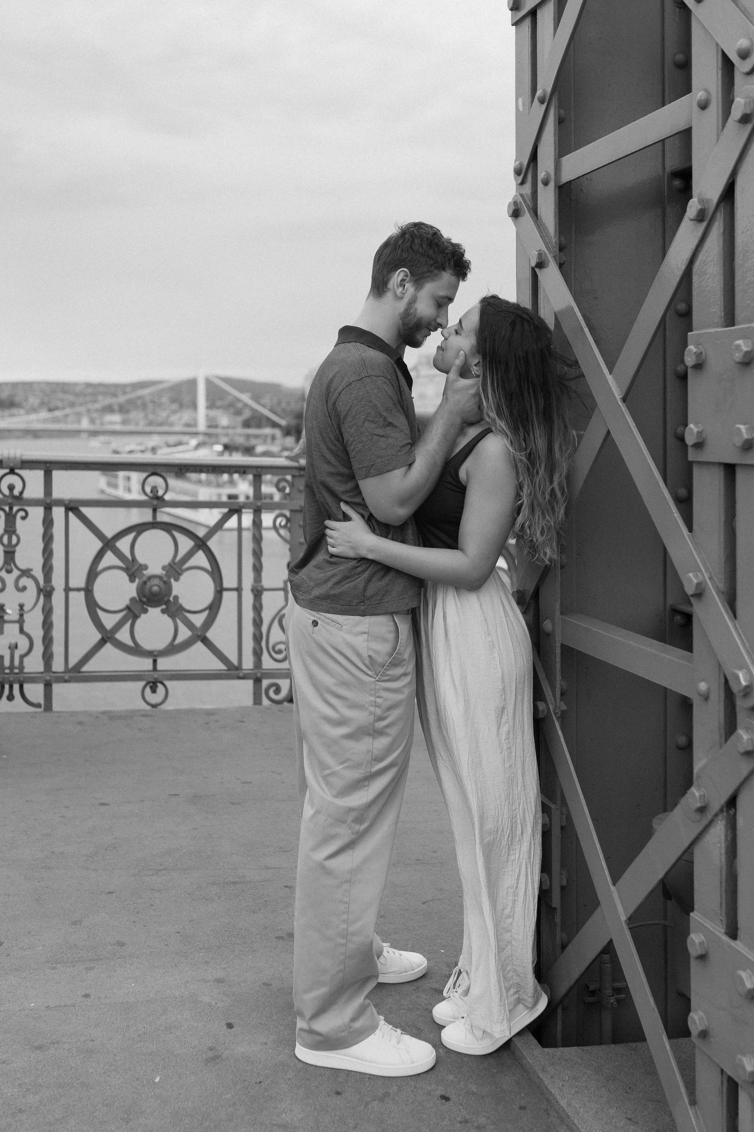 A black and white photo of a couple standing close to each other on a bridge, with the man holding the woman's face as they about to kiss. The woman is leaning against a large industrial structure, and there is a view of a bridge and landscape in the